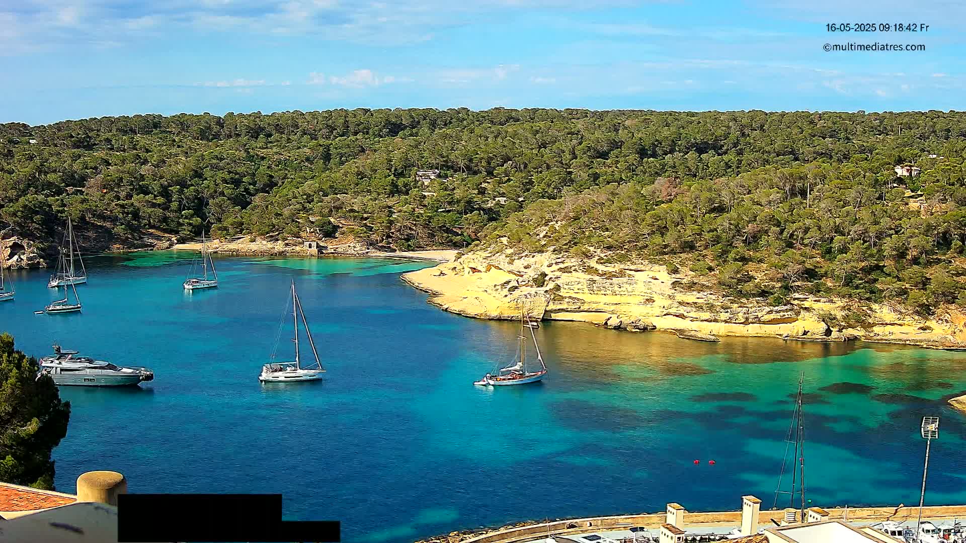 Several sailboats and a motor yacht are anchored in a calm, turquoise cove surrounded by lush green hills under a mostly sunny sky.