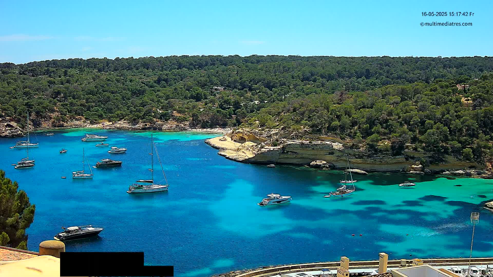 A cove of bright turquoise water is filled with sailboats and motorboats, surrounded by a lush green hillside under a clear blue sky.
