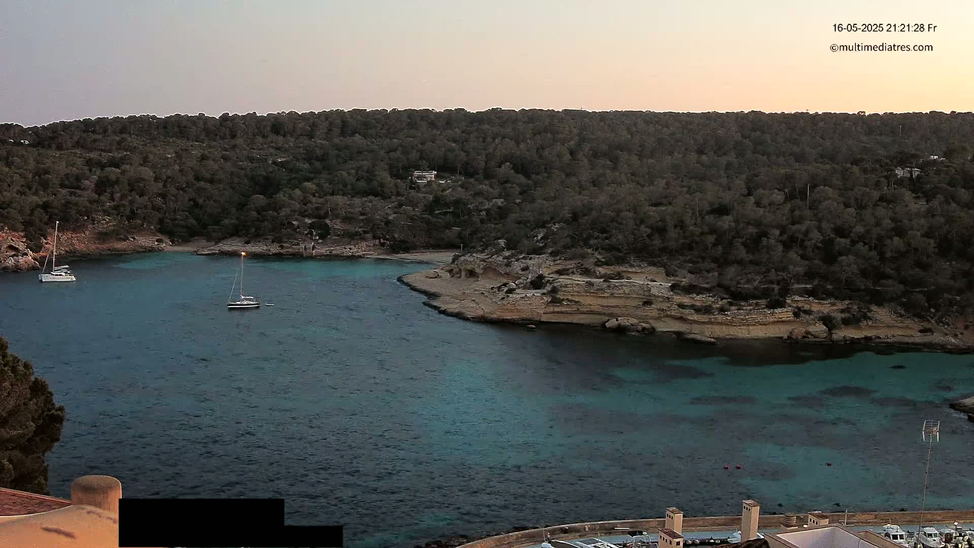 Two sailboats are anchored in a calm, teal-colored cove surrounded by dark green trees and light brown, rocky shores under a dusky sky.