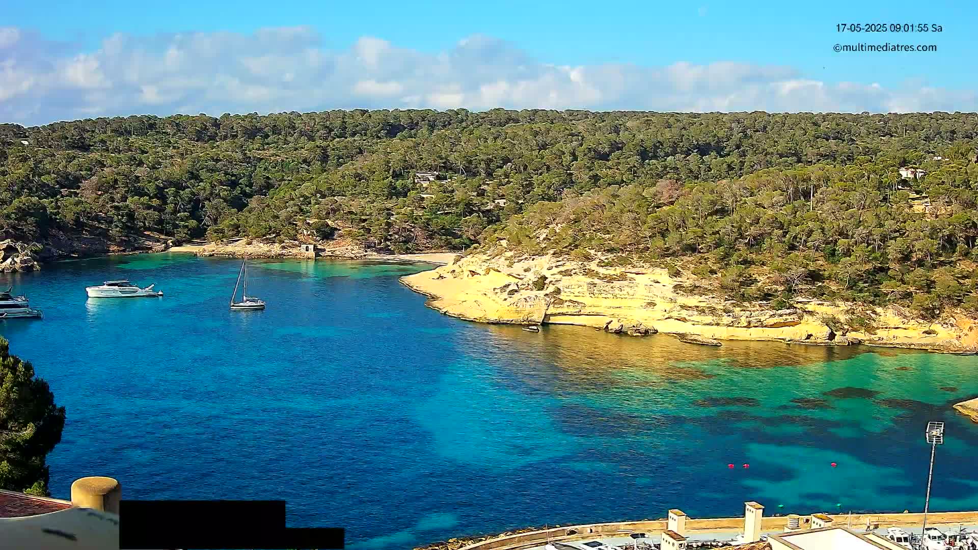 A tranquil, sunny cove with clear turquoise water, several boats, and a surrounding landscape of lush green trees and light-colored cliffs.