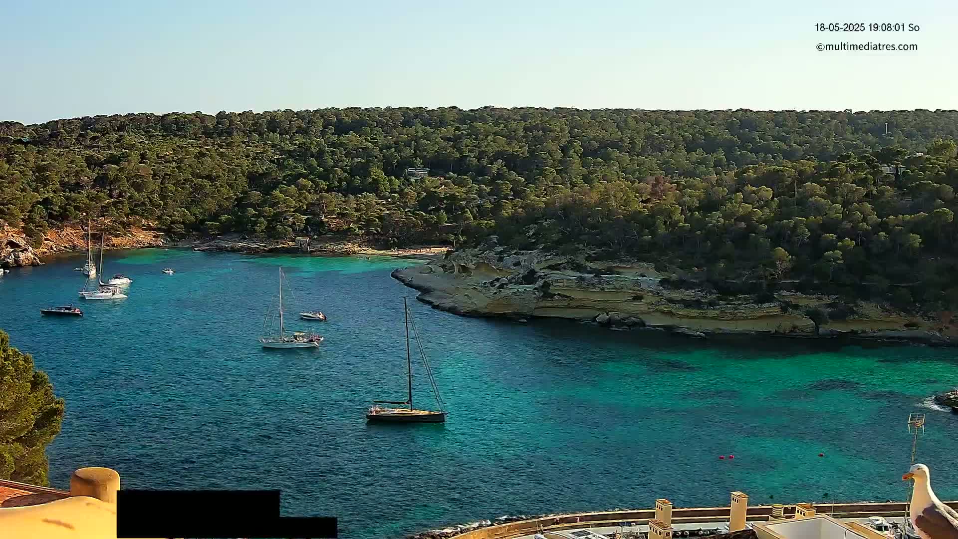 A tranquil, sunny cove with turquoise water is surrounded by lush green hillsides, and several sailboats are anchored in the calm bay.