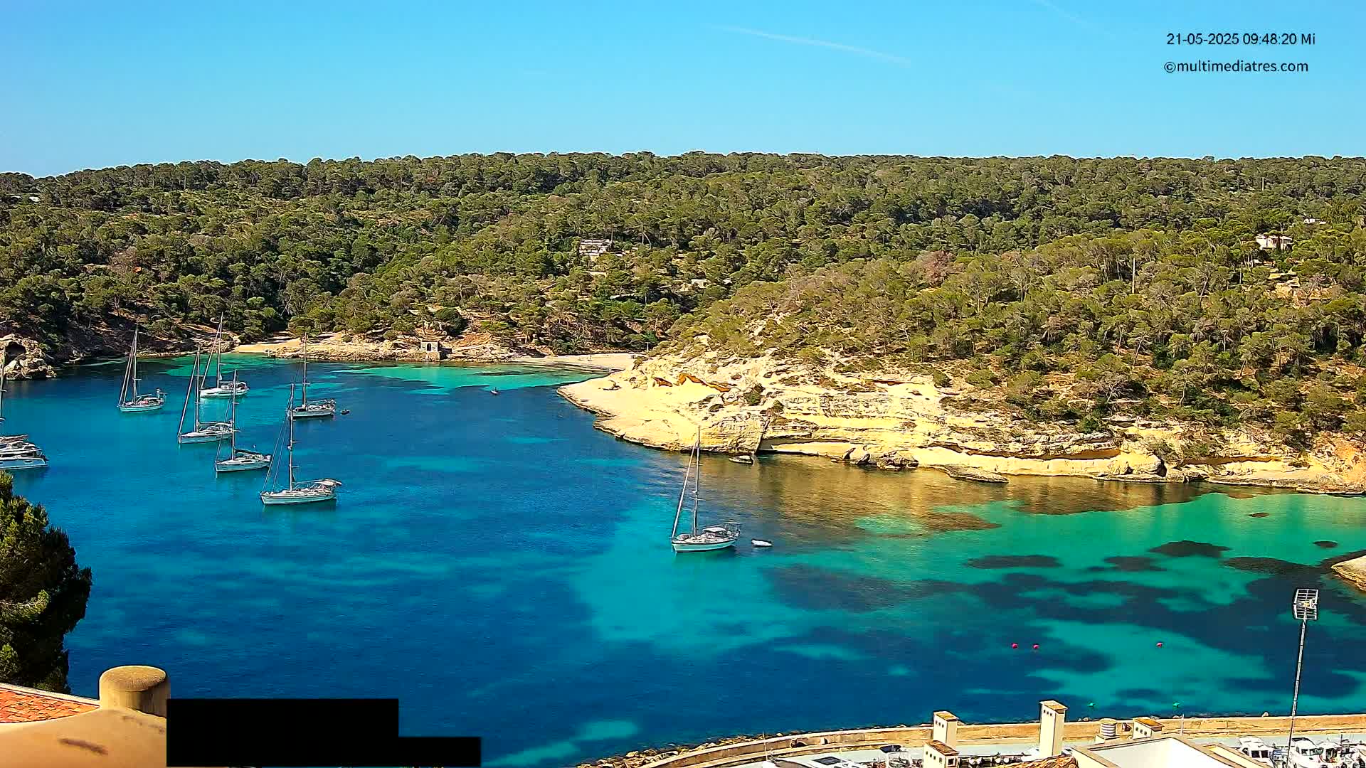 Several sailboats are anchored in a calm, clear turquoise cove surrounded by a lush green hillside under a bright blue sky.