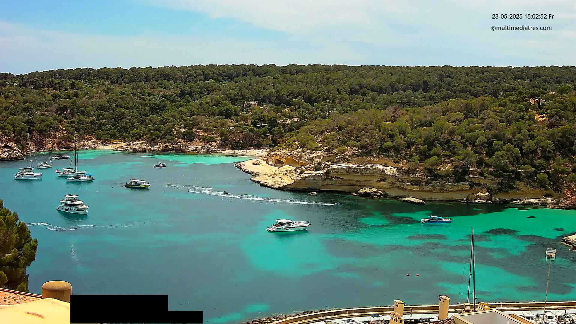 Several boats are anchored in a calm, turquoise cove surrounded by a lush, green hillside under a sunny sky.