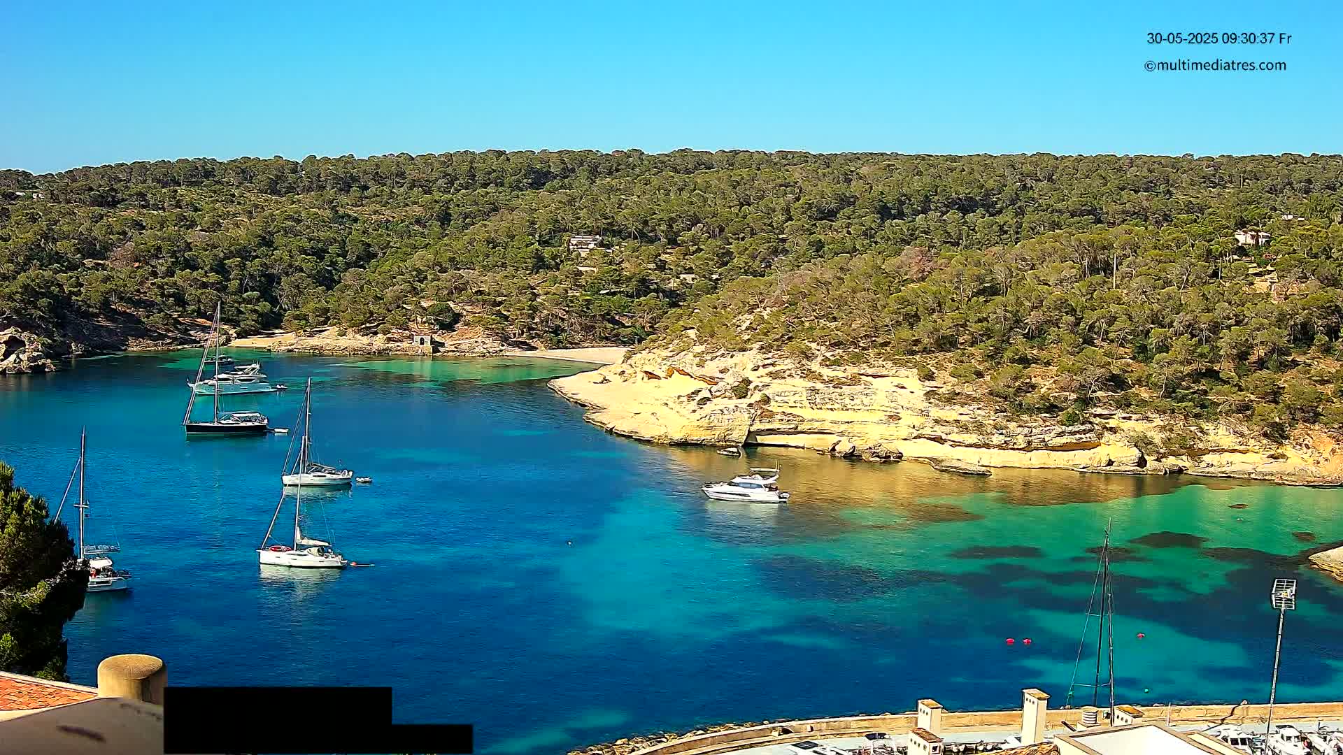 A secluded, bright blue cove surrounded by lush green hillsides and several sailboats and motorboats, under a clear sunny sky.