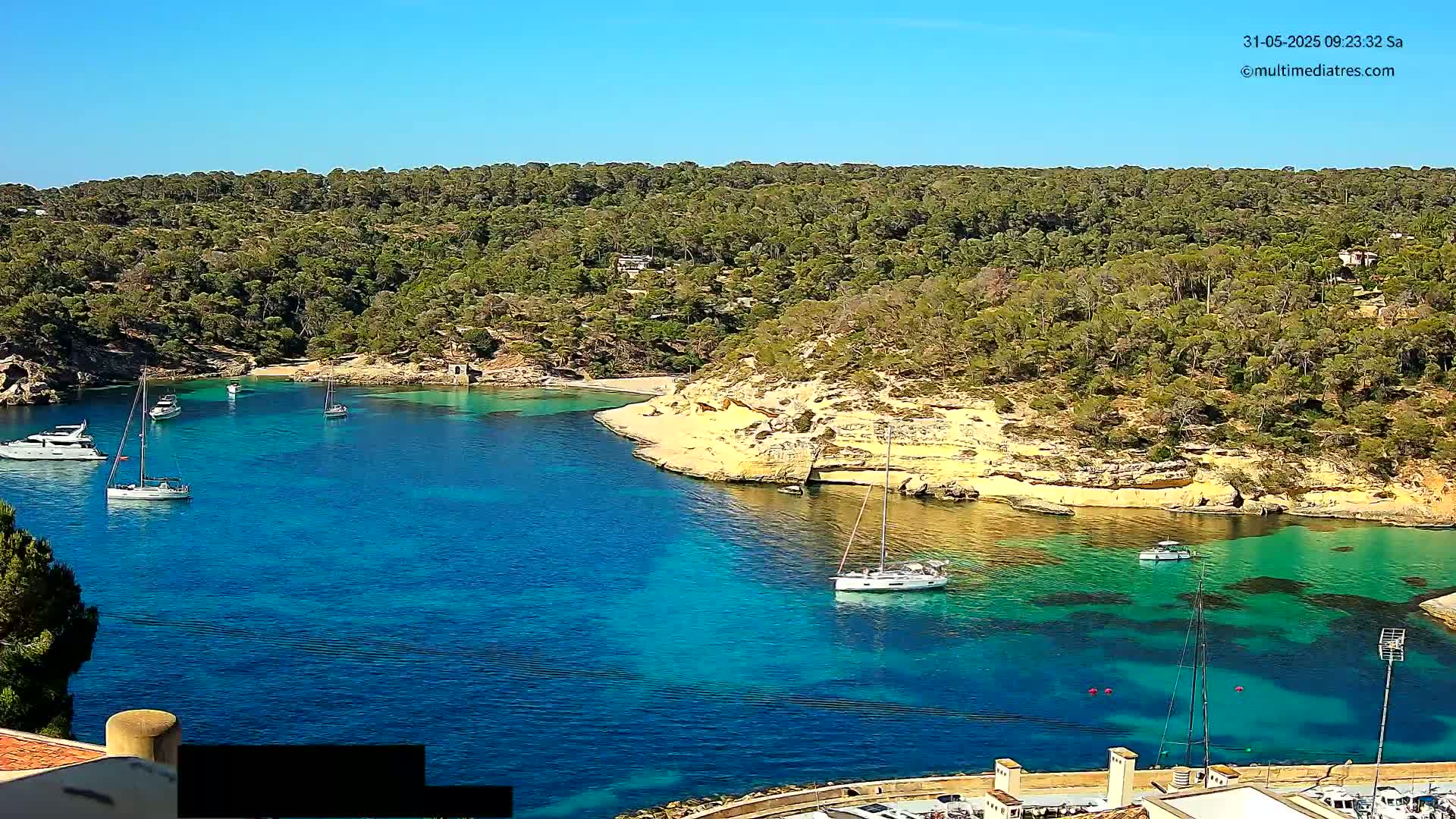 A secluded cove with crystal-clear turquoise water is surrounded by lush green vegetation under a bright, sunny sky, and several sailboats are moored in the calm bay.