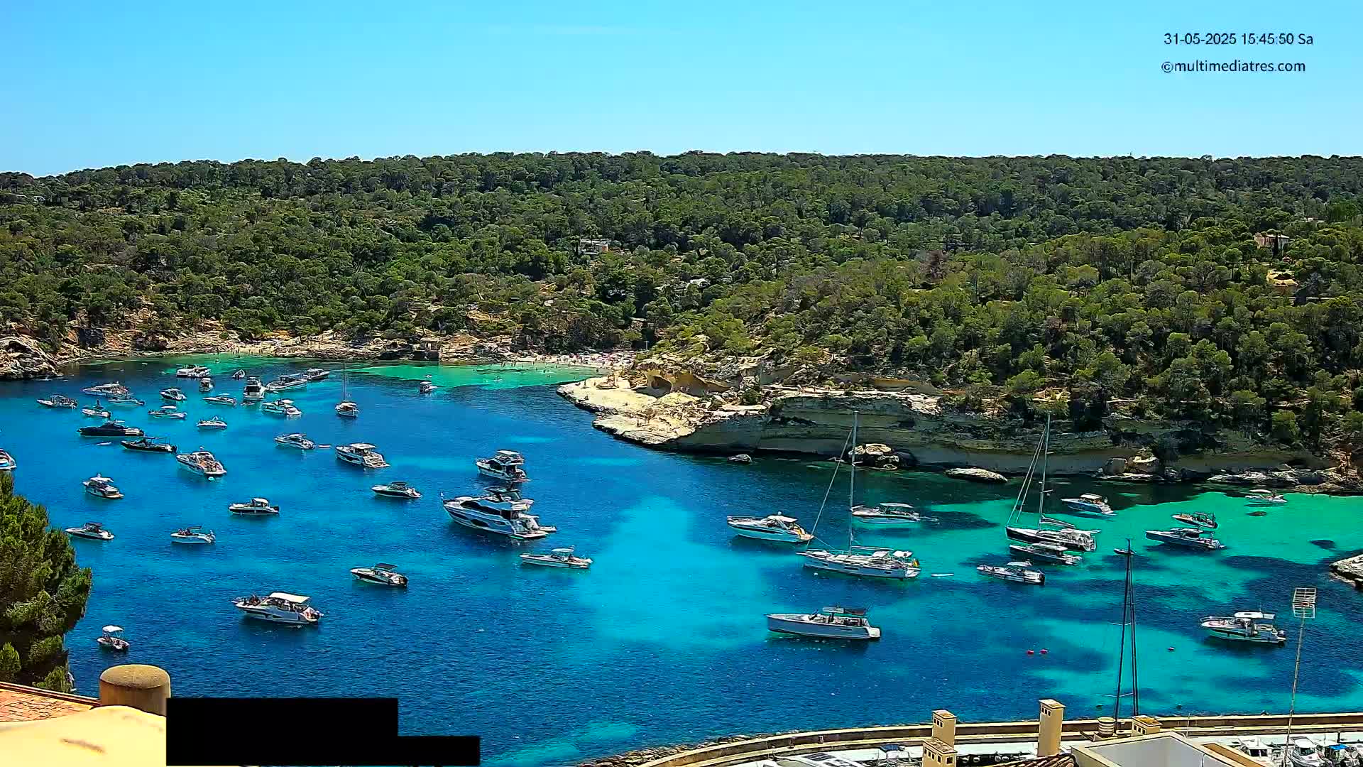 Numerous boats are anchored in a clear, turquoise cove surrounded by lush green hills under a bright, sunny sky.