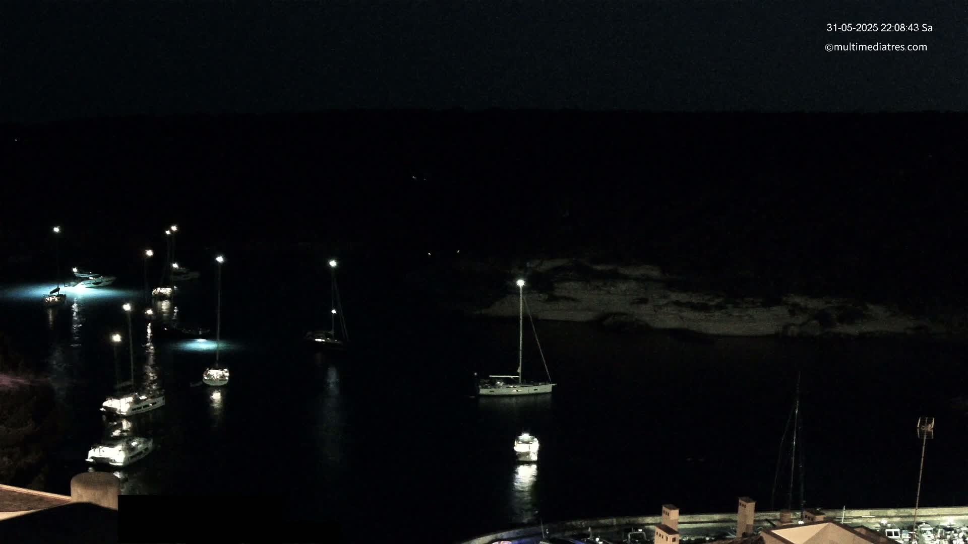 Several sailboats are moored in a dark, calm harbor at night, illuminated by individual lights.