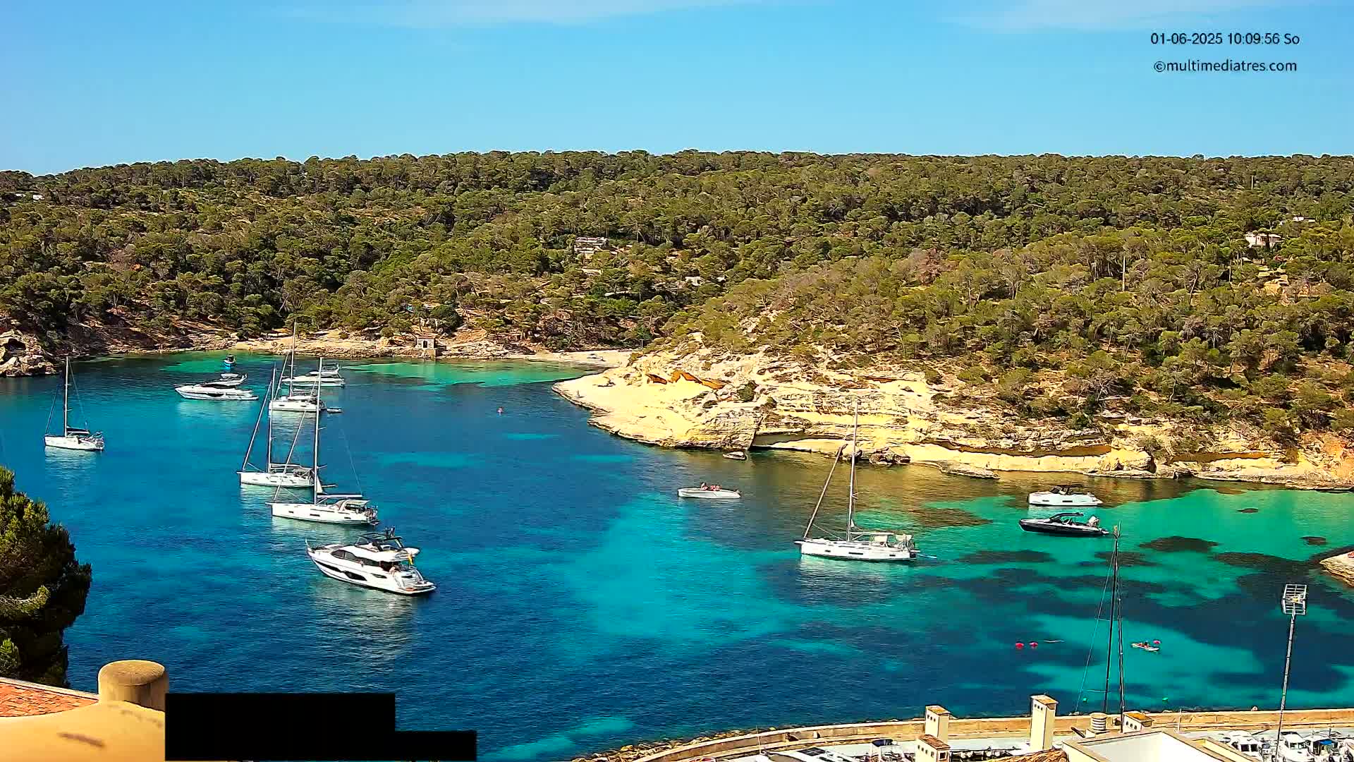 A tranquil cove with crystal-clear turquoise water, surrounded by lush green hills, is dotted with several sailboats and motorboats under a sunny sky.