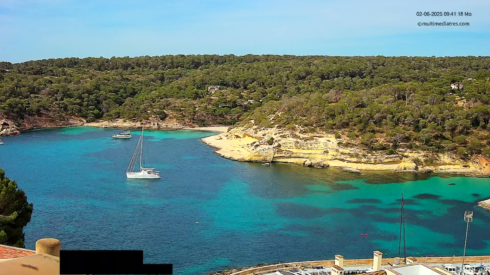 A tranquil cove with clear turquoise water, surrounded by lush green vegetation and light-colored cliffs under a sunny sky, features several sailboats and motorboats.