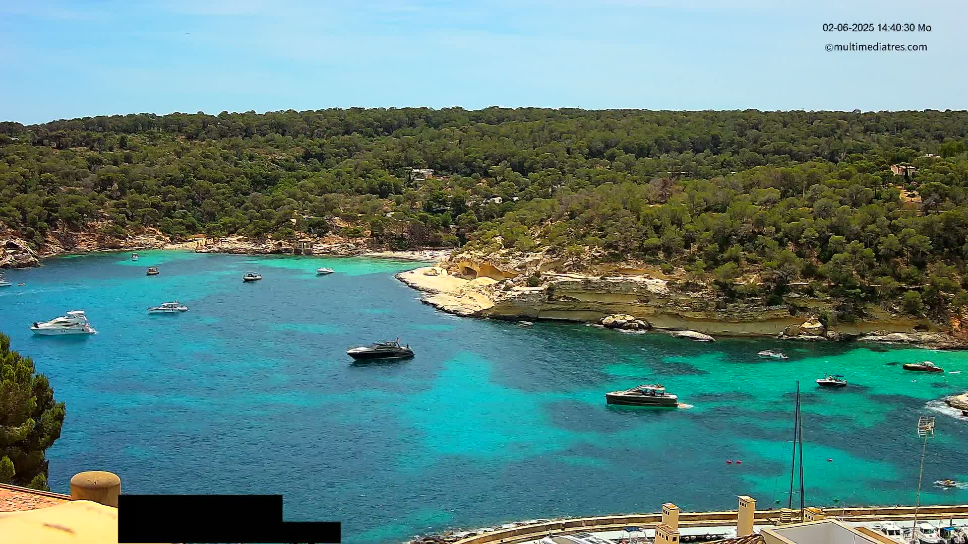A picturesque cove with clear, turquoise water, numerous boats, and a sandy beach surrounded by lush green hills under a sunny sky.