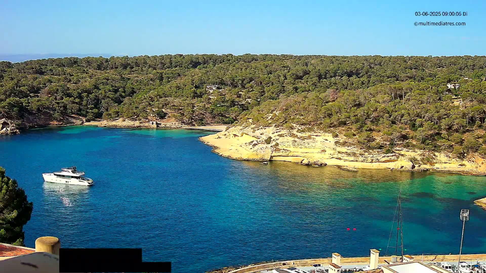 A yacht is anchored in a calm, clear blue bay surrounded by a lush green forest and pale yellow-tan cliffs under a bright sunny sky.