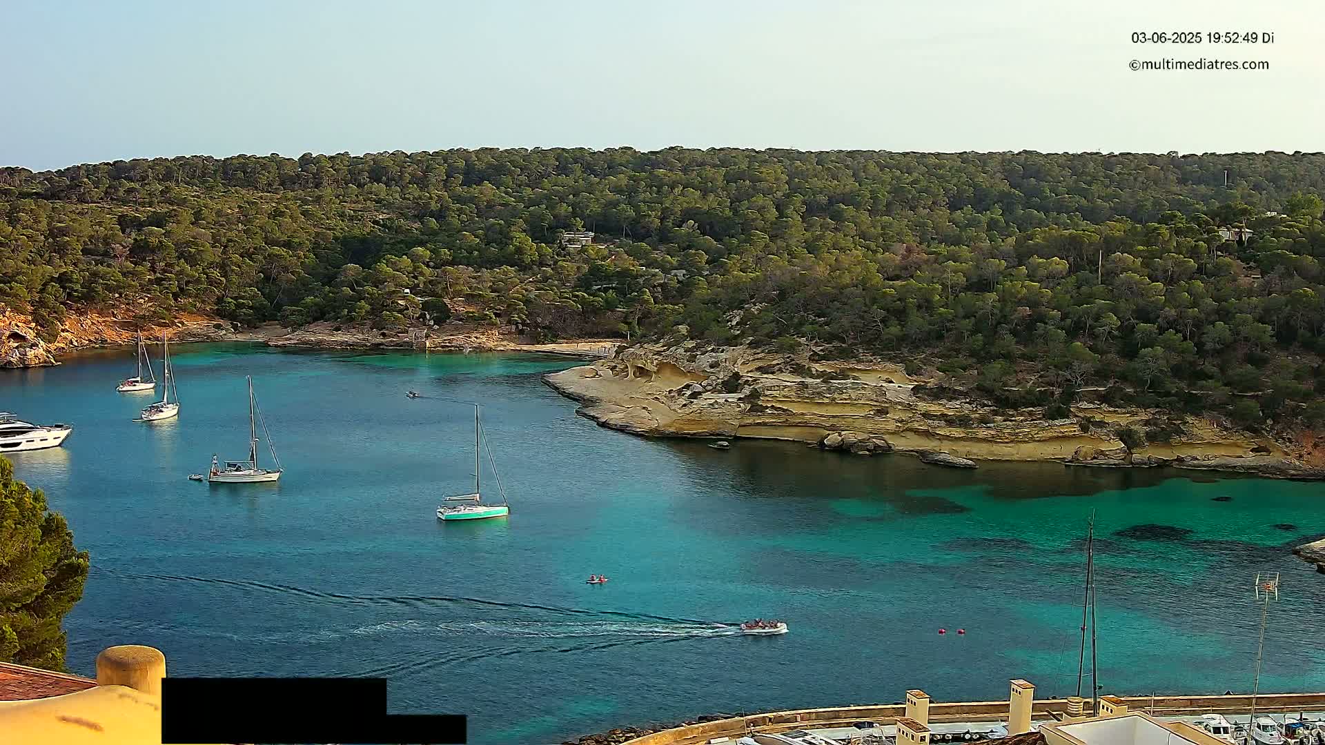 Several sailboats are anchored in a calm, turquoise cove surrounded by lush green hills under a clear sky.