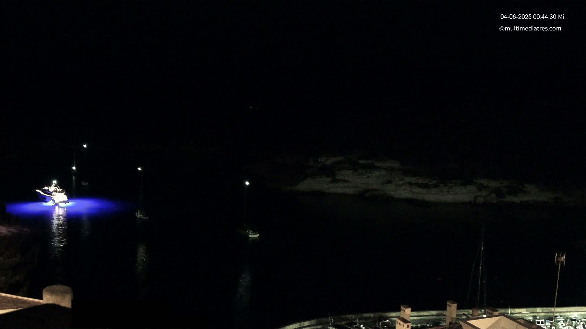 A nighttime, dark scene shows a body of water with a few boats, some illuminated with blue lights, near a dark shoreline.