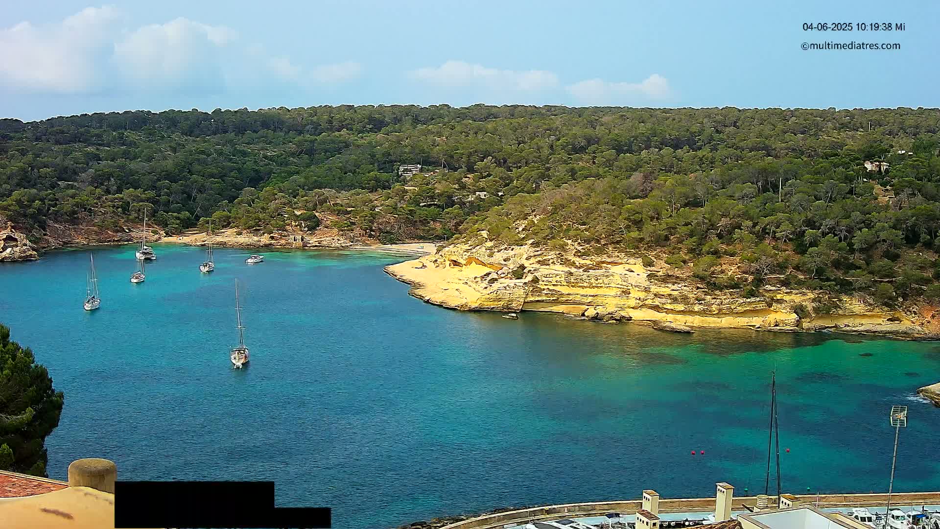 A picturesque cove with several sailboats is nestled within a lush, green hillside under a partly cloudy sky.