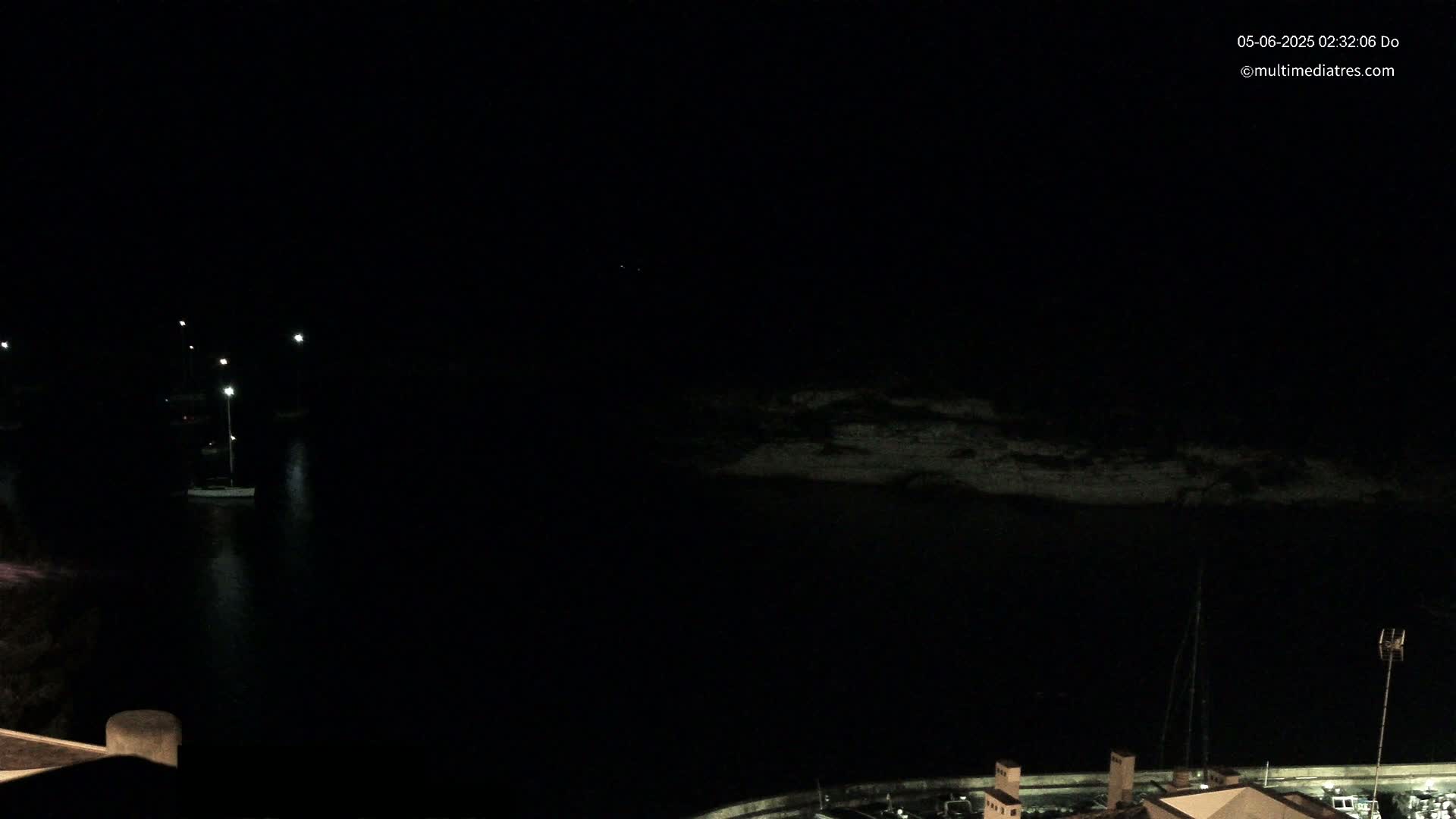 A nighttime view of a dark body of water with a sailboat and dimly lit shoreline.