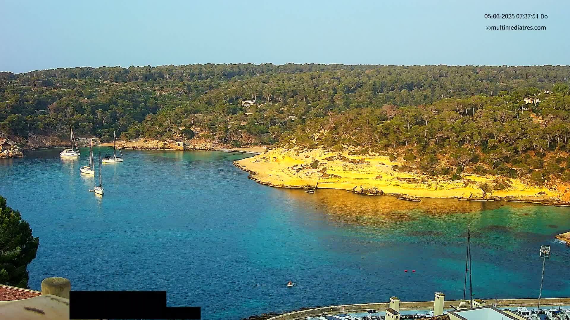 Several sailboats are anchored in a calm, clear turquoise bay surrounded by a lush green, hilly coastline under a bright, sunny sky.