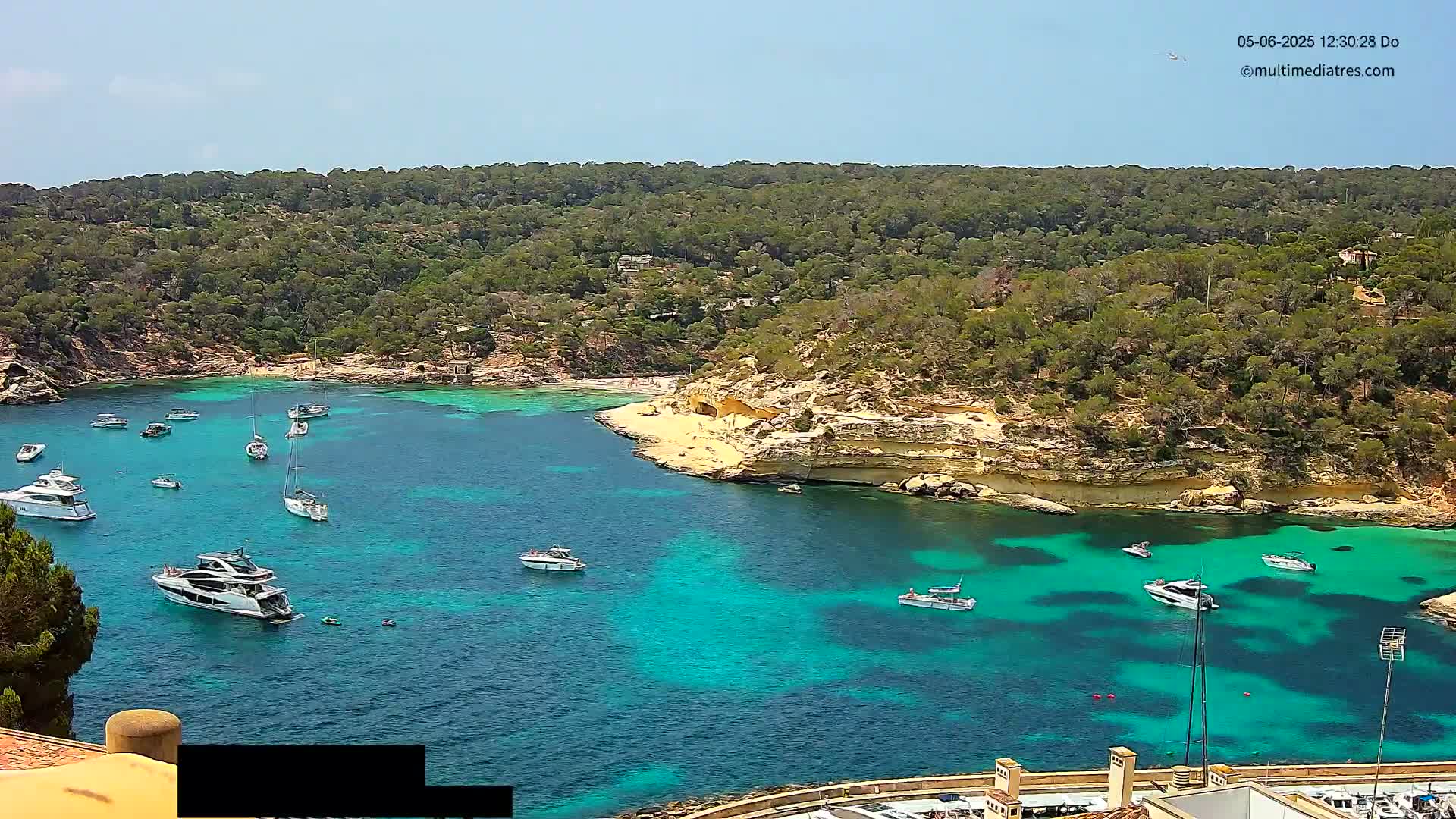 A secluded cove with turquoise water is surrounded by green hills under a clear, sunny sky, and numerous boats are anchored in the bay.