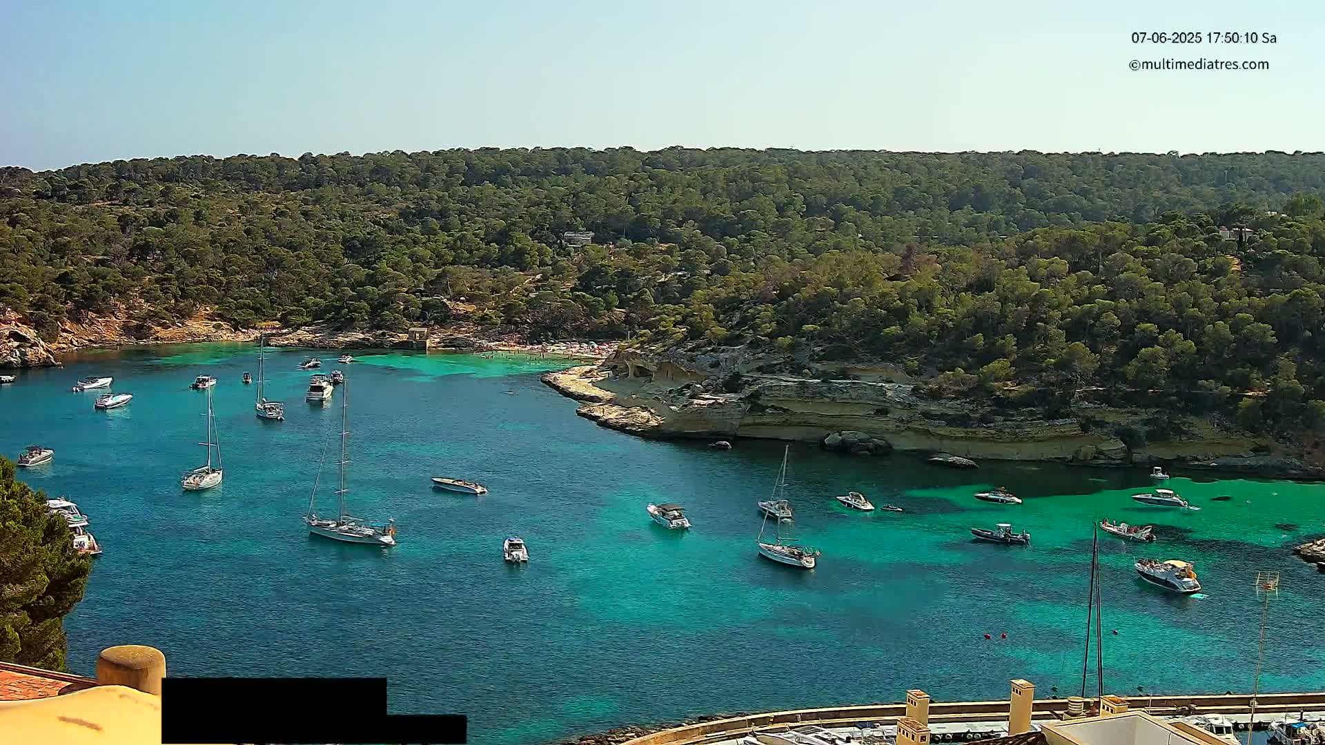 A tranquil bay with clear turquoise water, numerous boats anchored, and a surrounding landscape of lush green trees under a sunny sky.