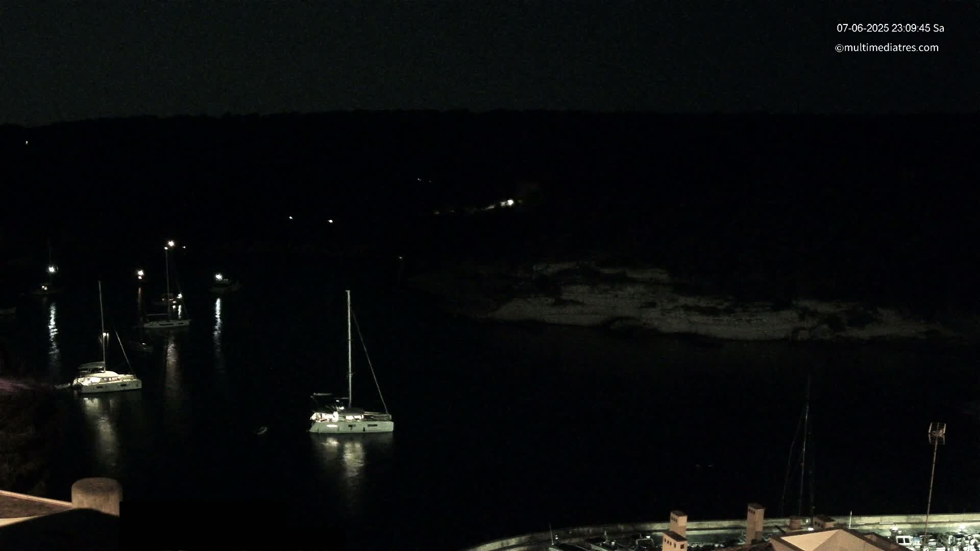 Several sailboats are moored in a dark, calm harbor at night, with a dark shoreline visible in the background.