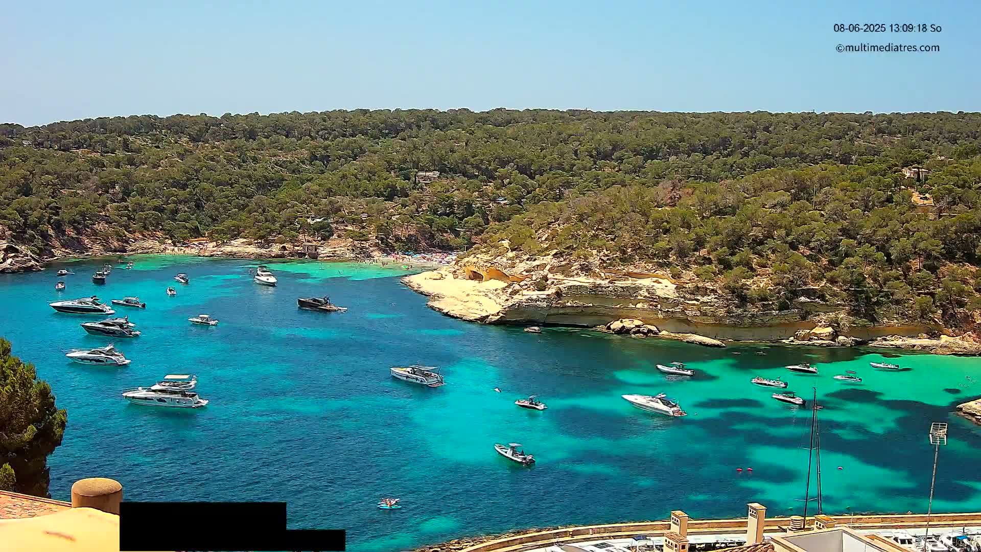 A sunny day reveals a secluded cove with turquoise water, numerous boats anchored, and a sandy beach surrounded by lush green hills.