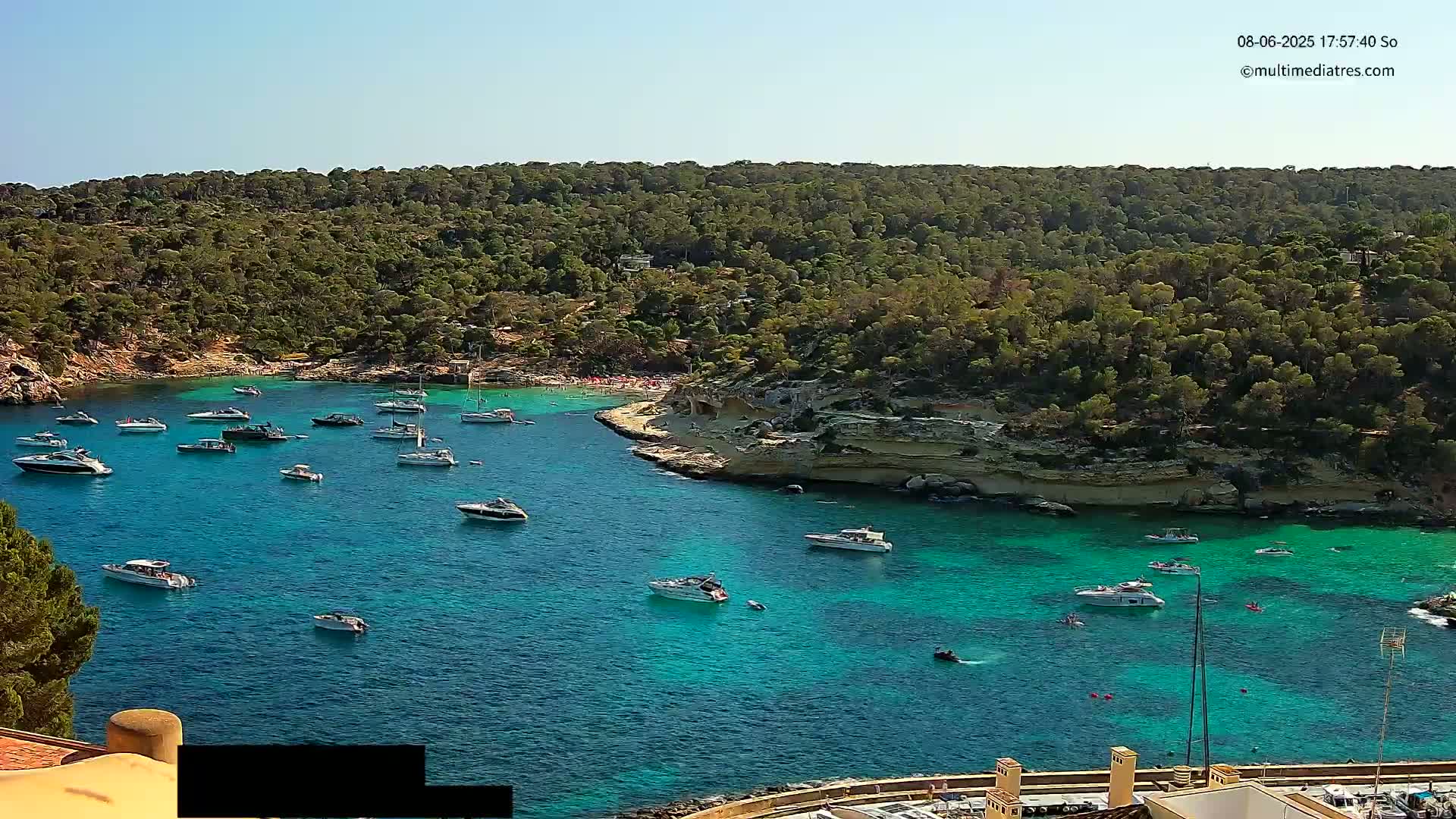 A secluded cove with turquoise water is filled with numerous boats, surrounded by lush green vegetation under a clear blue sky.