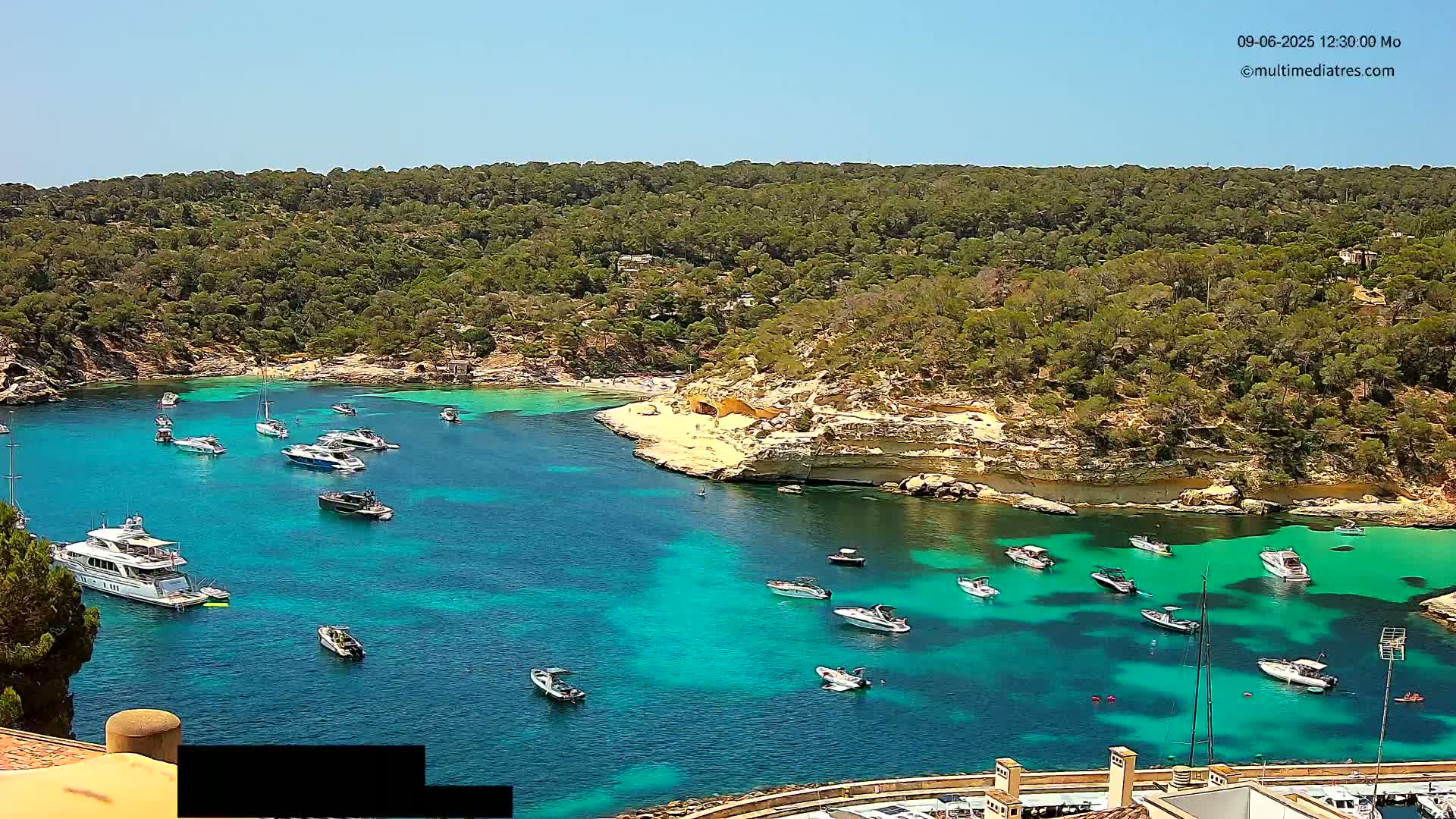 A secluded cove with clear turquoise water is surrounded by lush green vegetation, and numerous boats are anchored in the calm bay under a sunny sky.