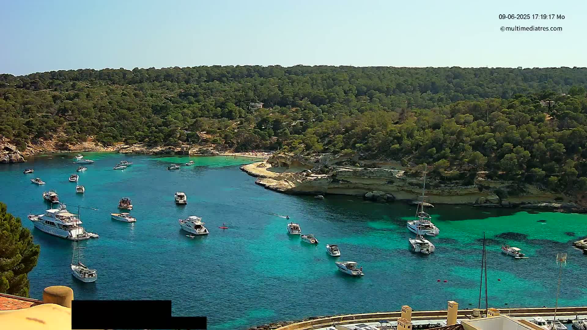 A secluded cove with turquoise water is filled with numerous boats and yachts, surrounded by lush green vegetation under a clear sky.