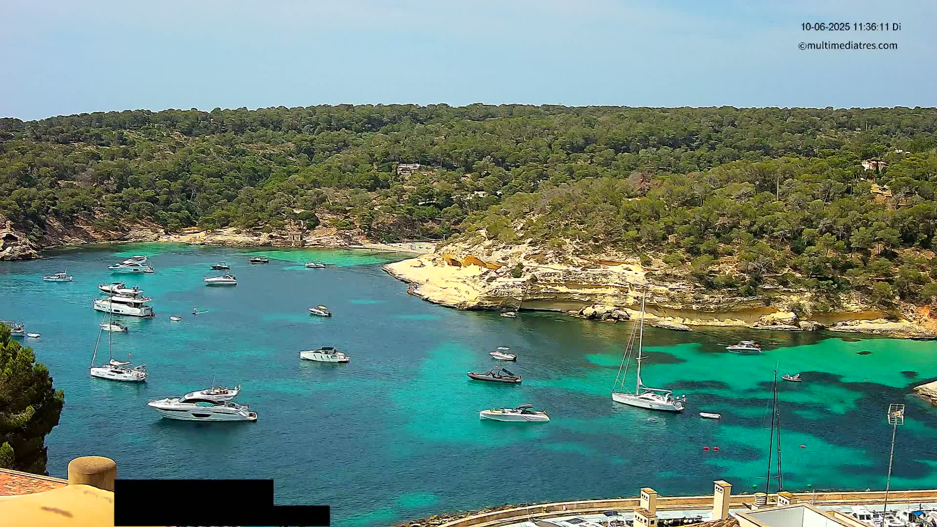 A secluded turquoise cove, surrounded by lush green vegetation, is filled with numerous boats on a sunny day.