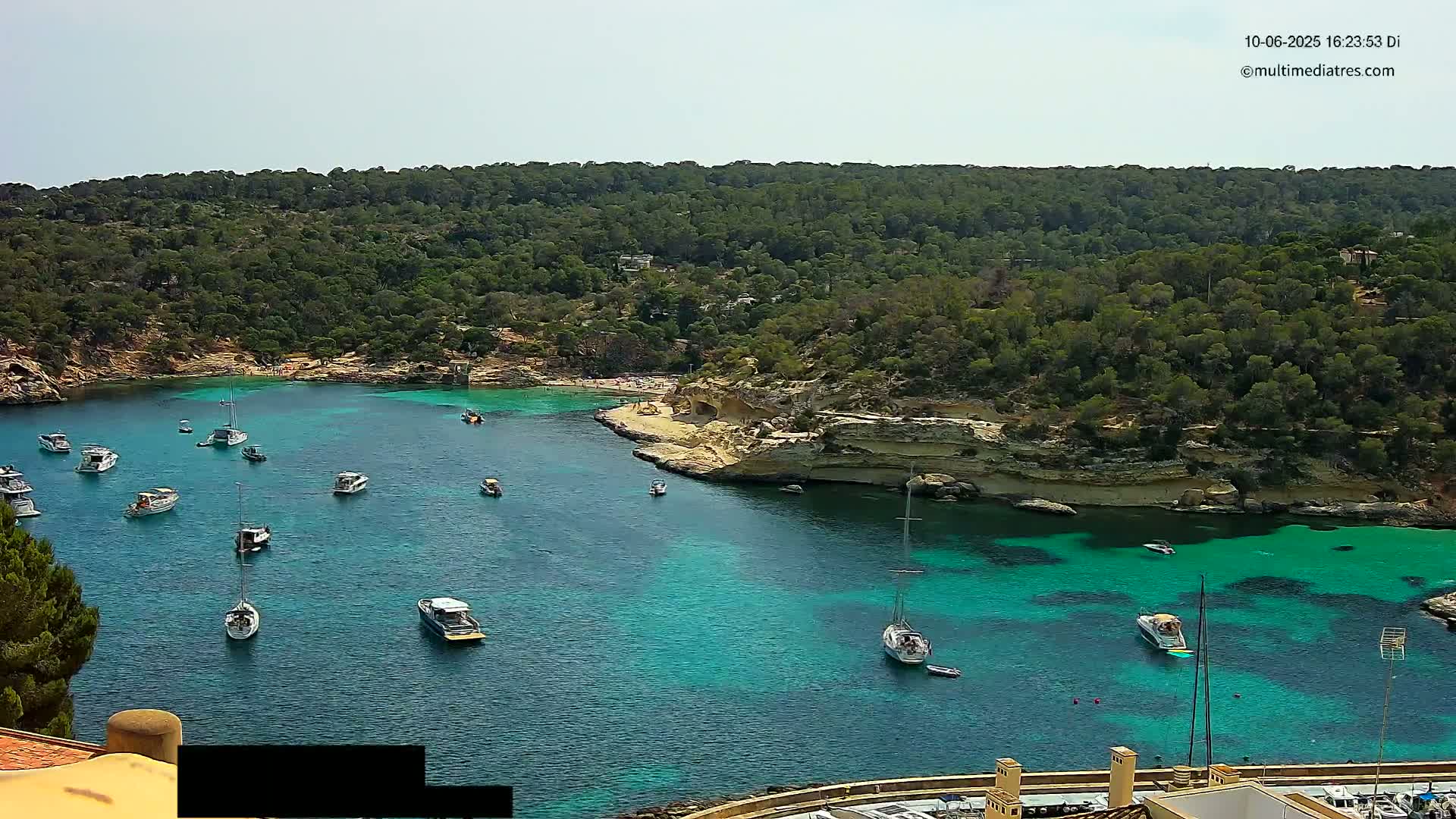 A tranquil cove with clear, turquoise water is filled with numerous boats, surrounded by lush green hills under a bright, sunny sky.