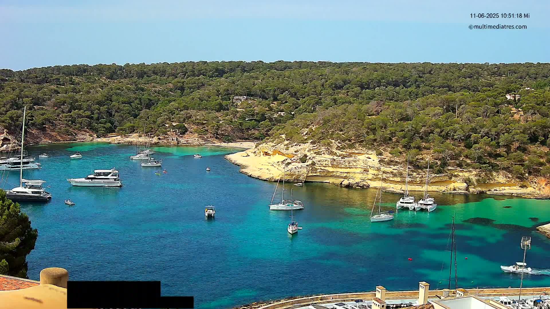 A secluded cove with turquoise water is surrounded by lush green hills under a clear blue sky, numerous sailboats and motorboats are anchored in the calm bay.