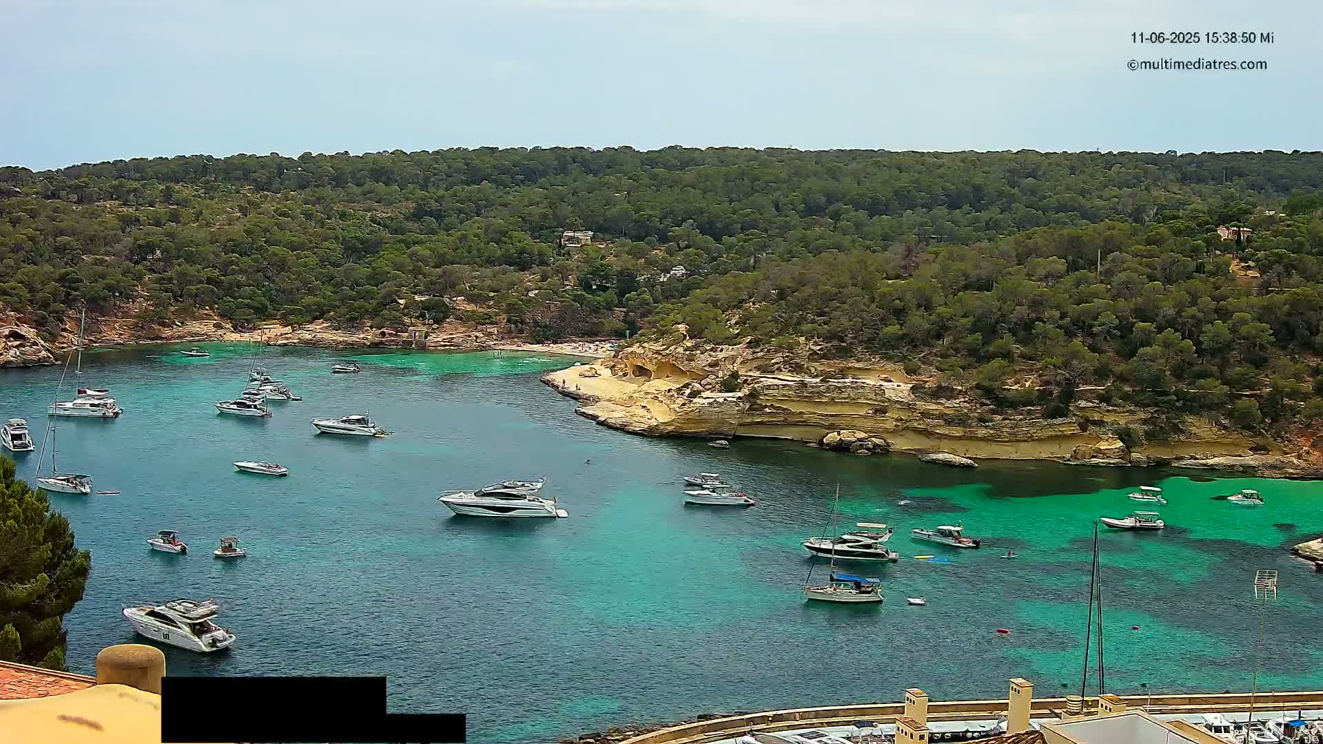 A calm, sunny day shows a cove of clear, teal water with numerous boats and yachts anchored, surrounded by lush green hillsides and tan-colored cliffs.