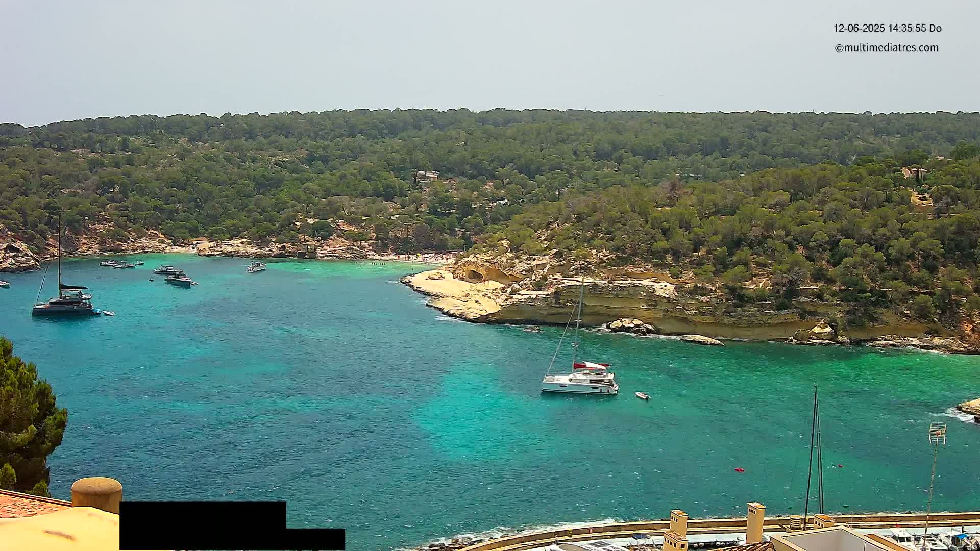 A secluded cove with turquoise water, surrounded by lush green hills and several boats anchored in the bay under sunny skies.