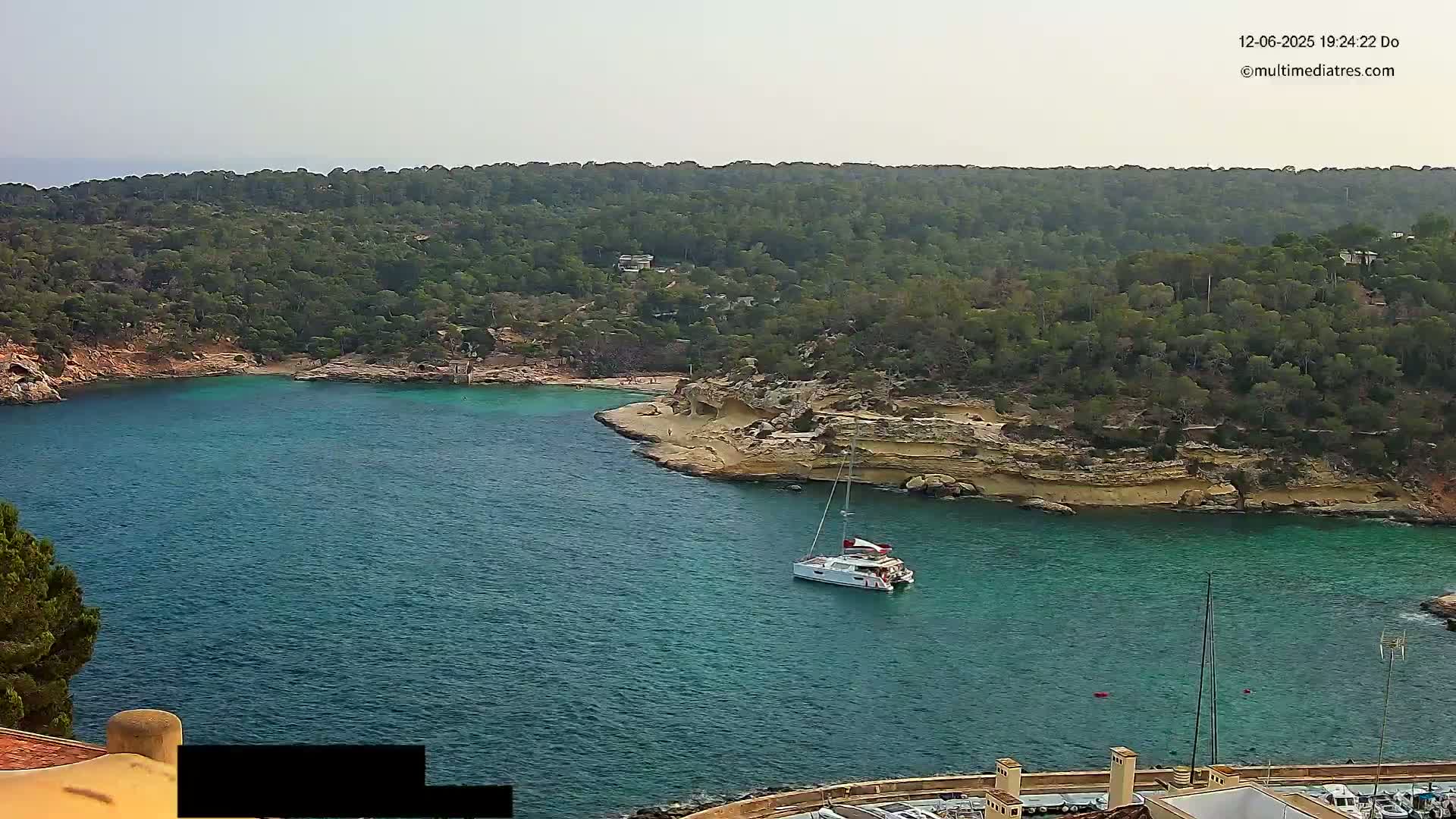 A sailboat is anchored in a calm, teal bay surrounded by a lush, green, hilly coastline under a hazy sky.