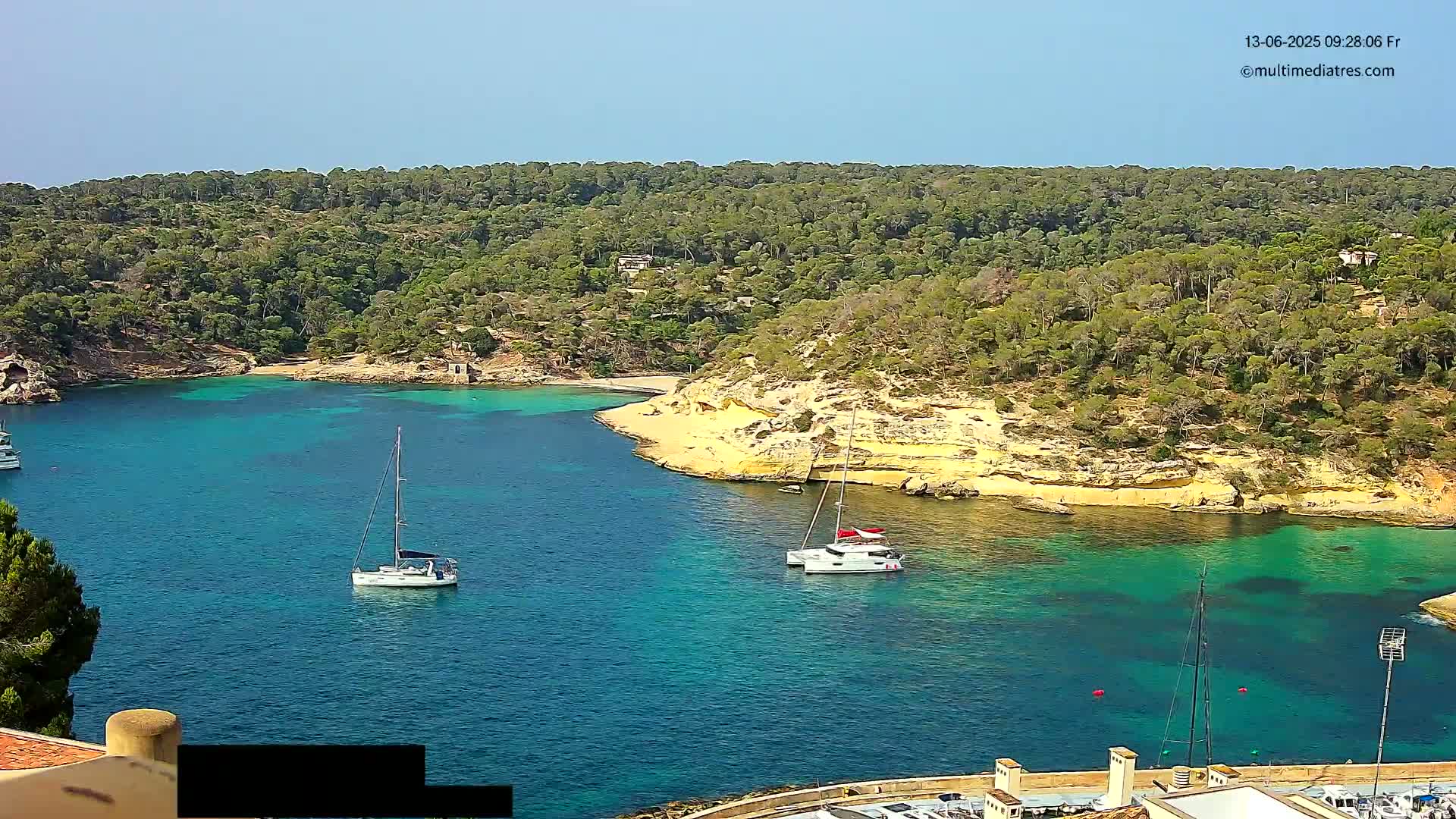 A sunny, clear day shows a secluded cove with turquoise water, surrounded by lush green trees and tan cliffs, where several sailboats are anchored.