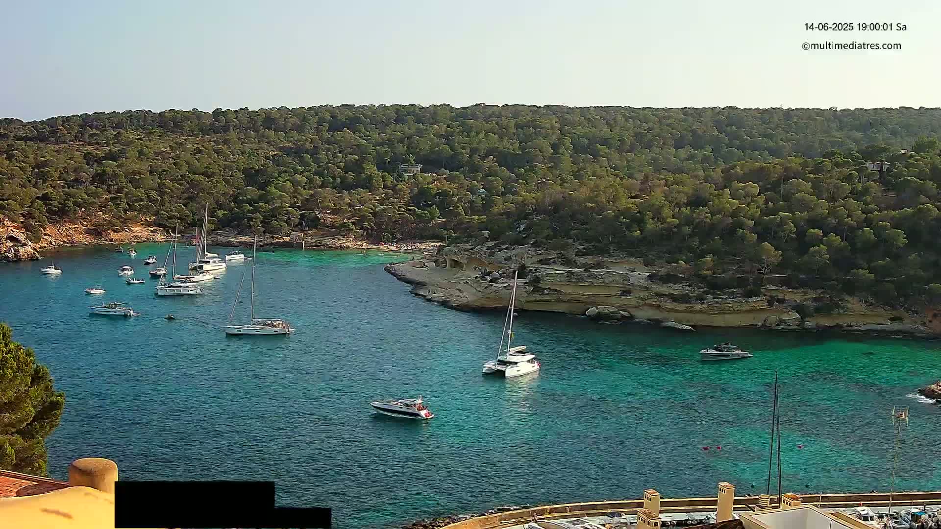 A calm, sunny day reveals a secluded cove filled with numerous sailboats and motorboats, nestled within a verdant, hilly landscape.