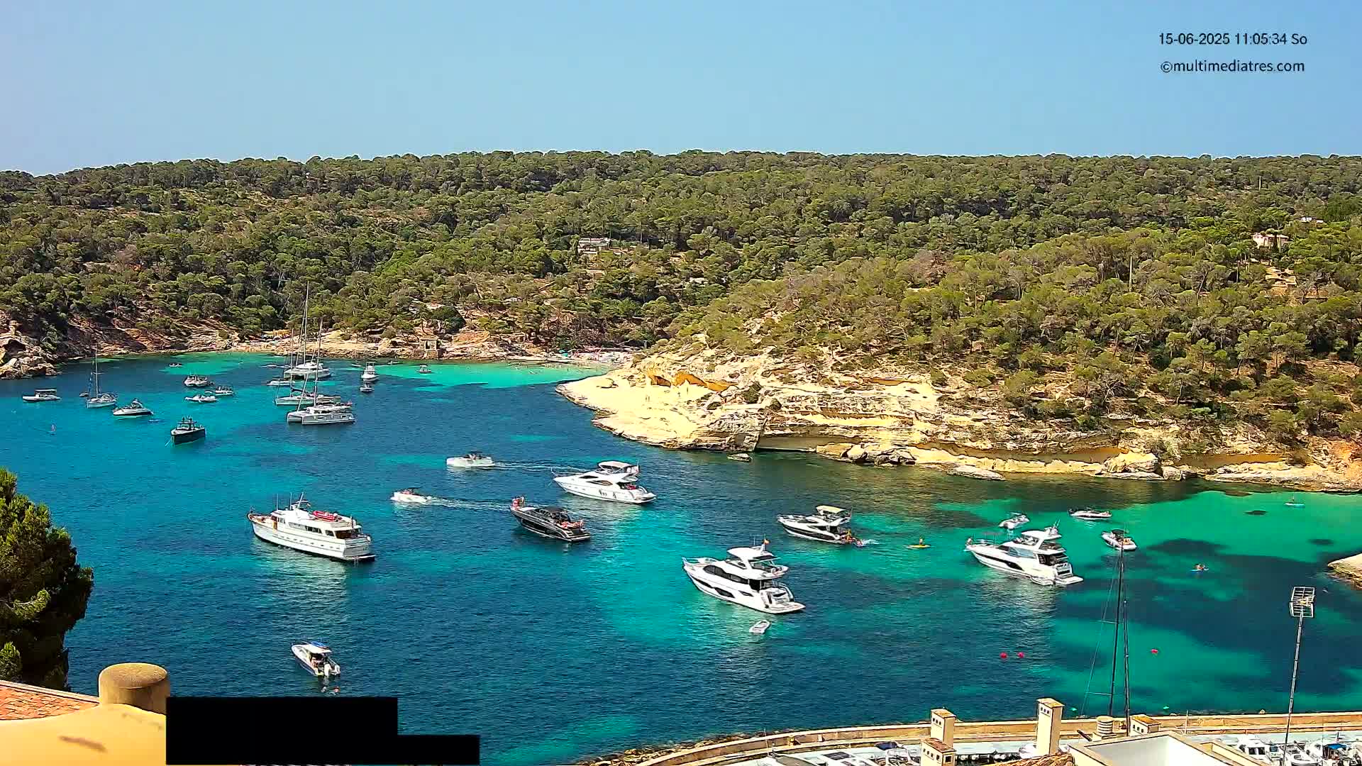 A calm, sunny day shows a cove filled with numerous boats of various sizes, anchored in bright turquoise water, surrounded by a lush green, rocky coastline.
