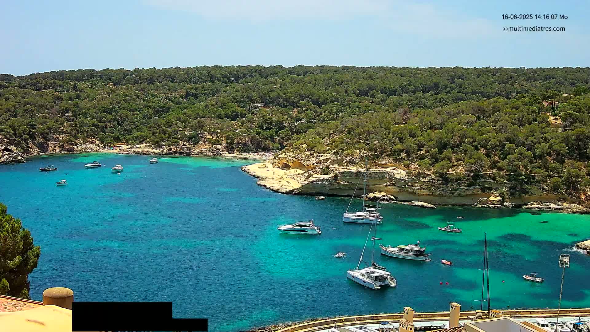 A tranquil, sunny cove with clear turquoise water is filled with several boats, anchored near a sandy beach surrounded by lush green vegetation.