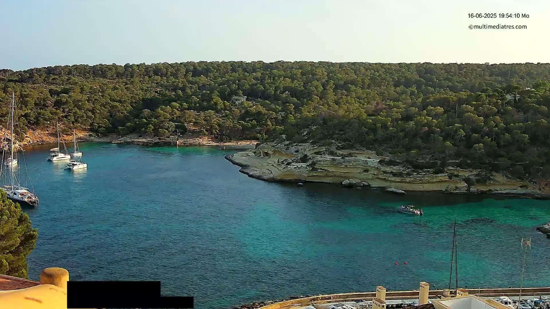 Several sailboats are anchored in a calm, clear turquoise cove surrounded by lush green hills under a clear sky.