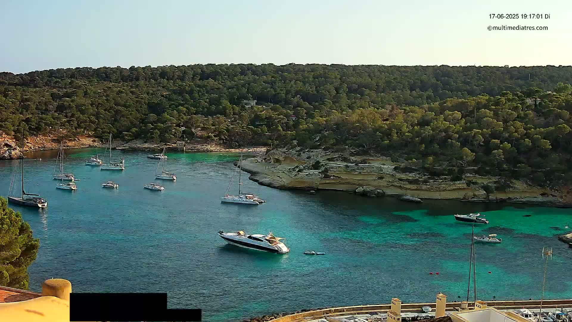 A tranquil, sunny cove filled with turquoise water and numerous sailboats and motorboats is surrounded by a lush, green hillside.