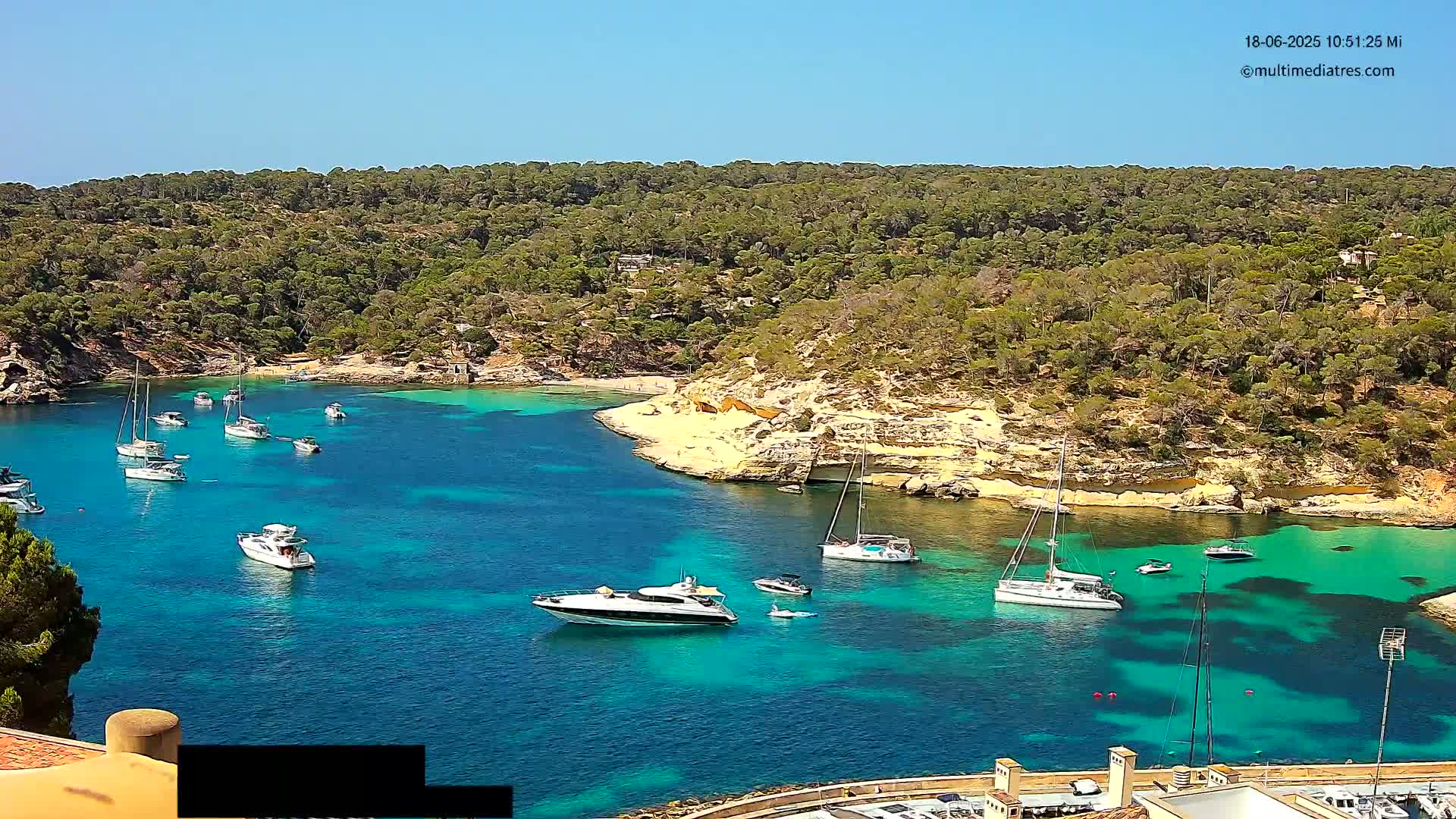 A secluded, turquoise cove is filled with sailboats and yachts, surrounded by a lush, green hillside under a clear, sunny sky.