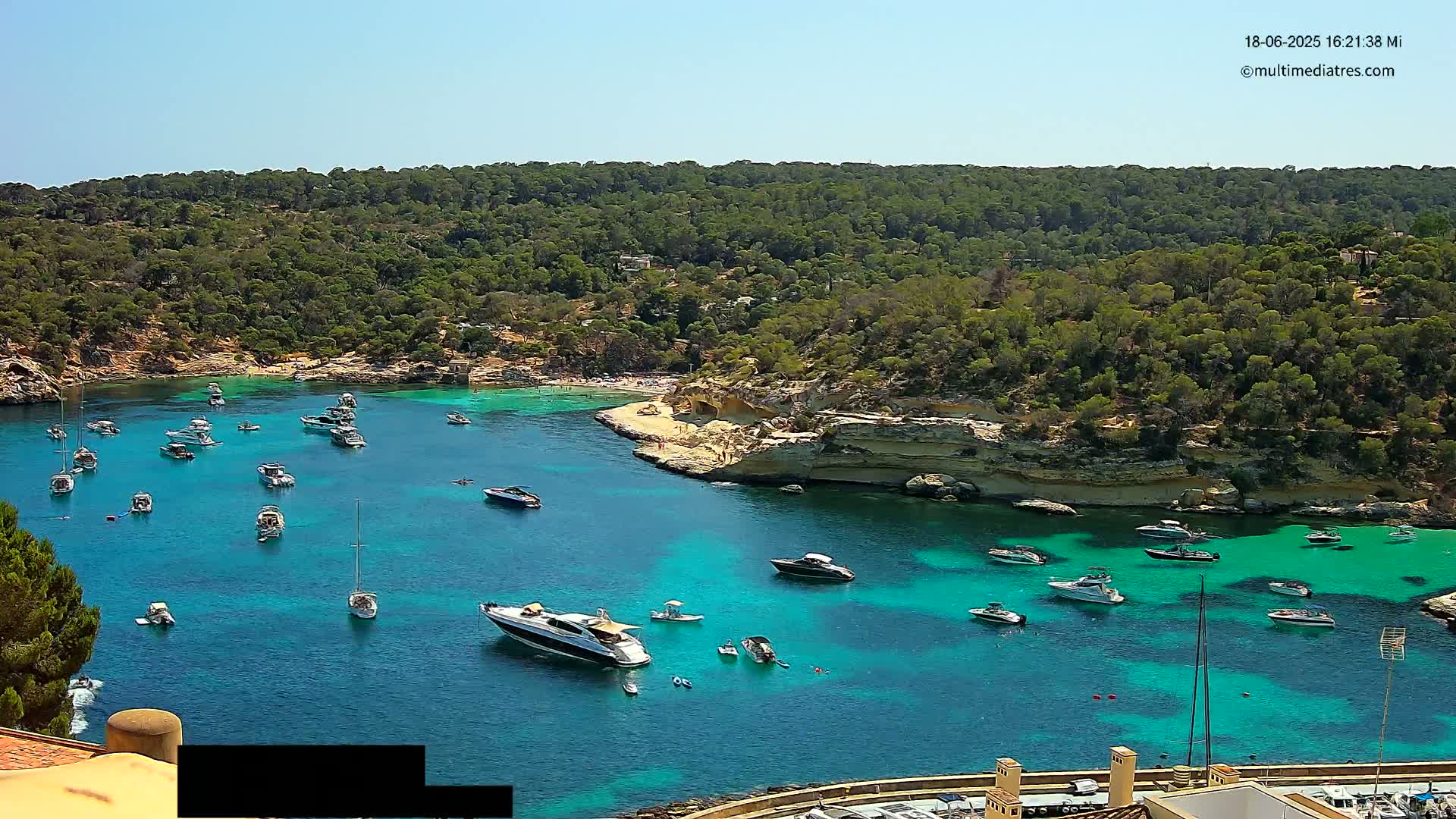 A secluded cove with crystal-clear turquoise water is filled with numerous boats and yachts, surrounded by lush green vegetation under a bright sunny sky.