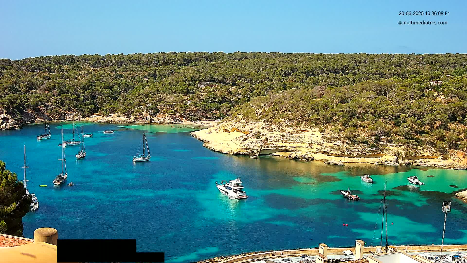 A secluded, turquoise cove surrounded by lush green hills and dotted with sailboats and a motor yacht under a clear blue sky.