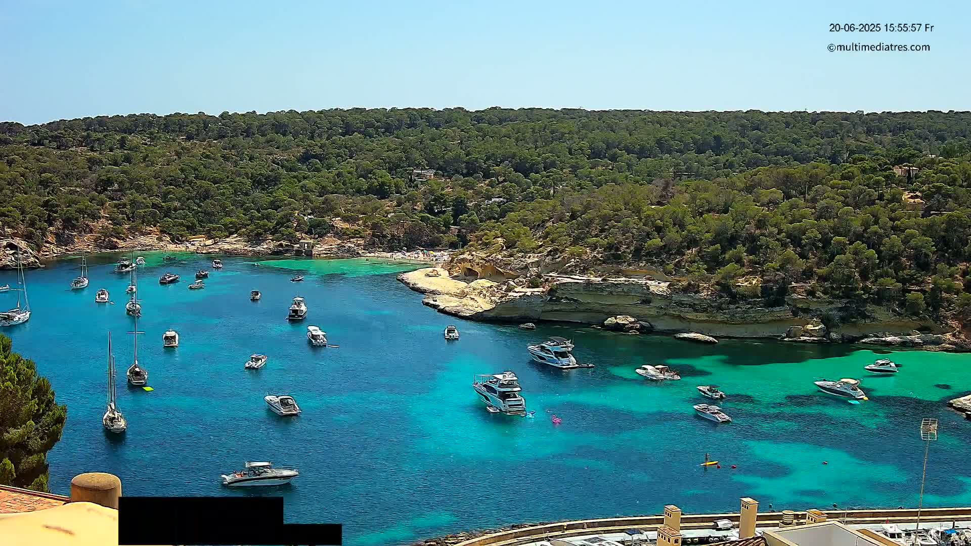A secluded cove with clear, turquoise water is filled with numerous boats and yachts, surrounded by lush green hills under a bright sunny sky.