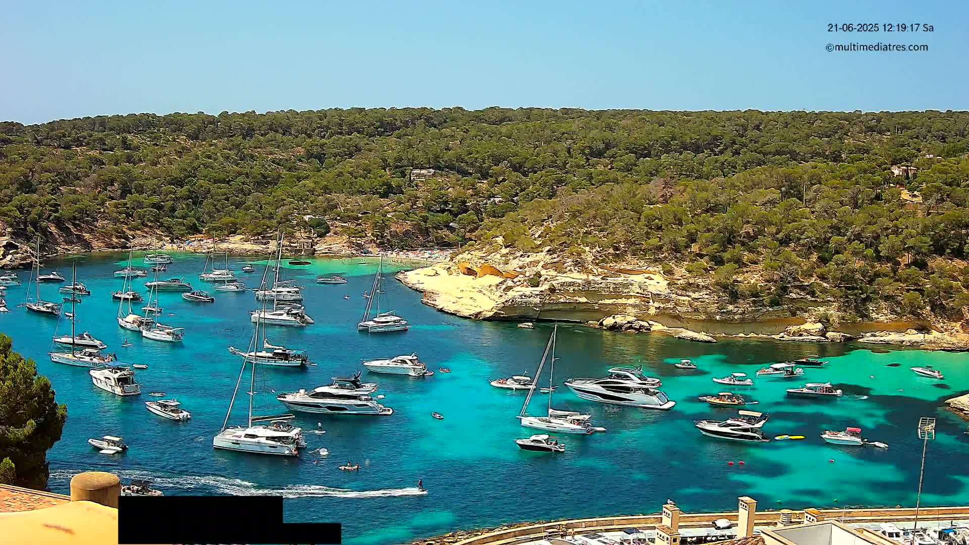 A sunny day reveals a secluded cove filled with numerous boats and yachts anchored in vibrant turquoise water, surrounded by lush green hills.