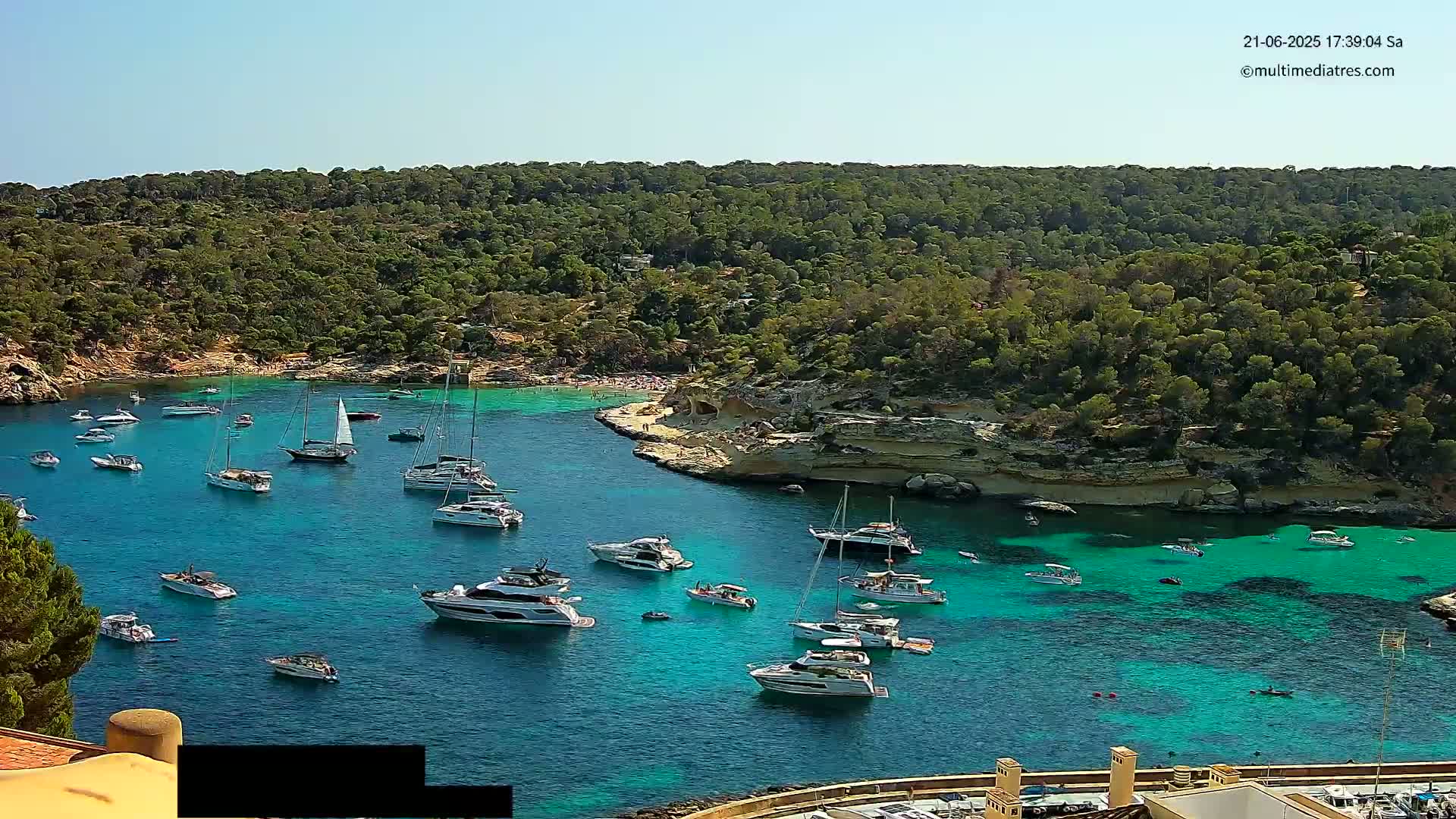A sunny day shows a calm, turquoise bay filled with numerous boats and yachts, surrounded by lush green hillsides.