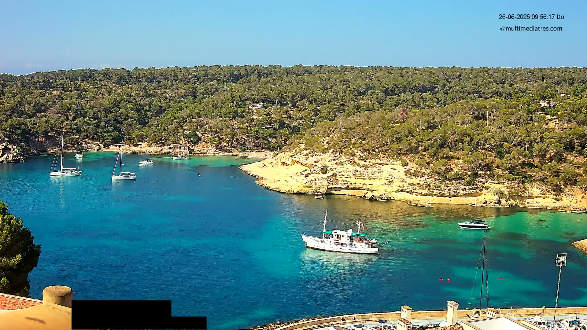 A tranquil bay of clear, turquoise water is surrounded by a lush, green, hilly coastline under a bright, sunny sky, with several sailboats and a larger motor vessel in the water.