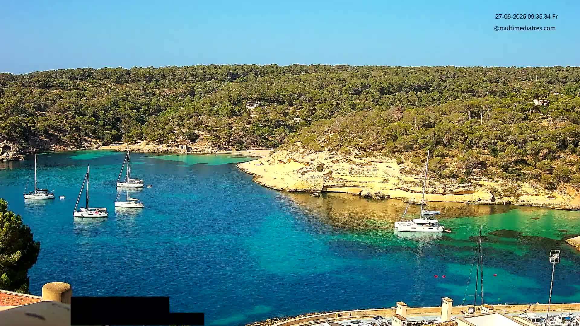 A secluded, sunny cove with turquoise water contains several sailboats moored near a sandy beach bordered by lush green trees.