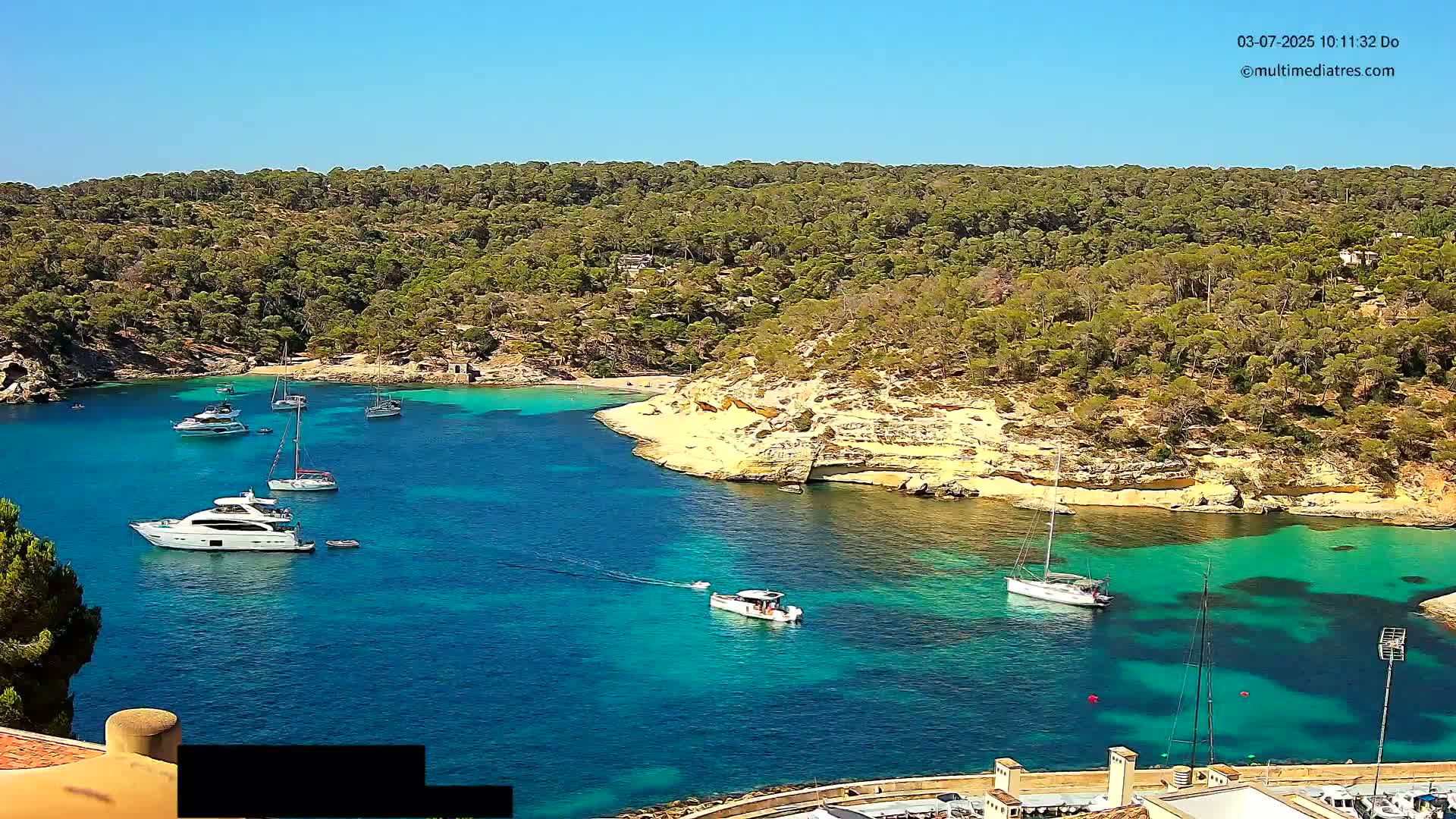 A calm, sunny day shows a secluded turquoise cove surrounded by lush green vegetation, with several sailboats and yachts anchored in the clear water.