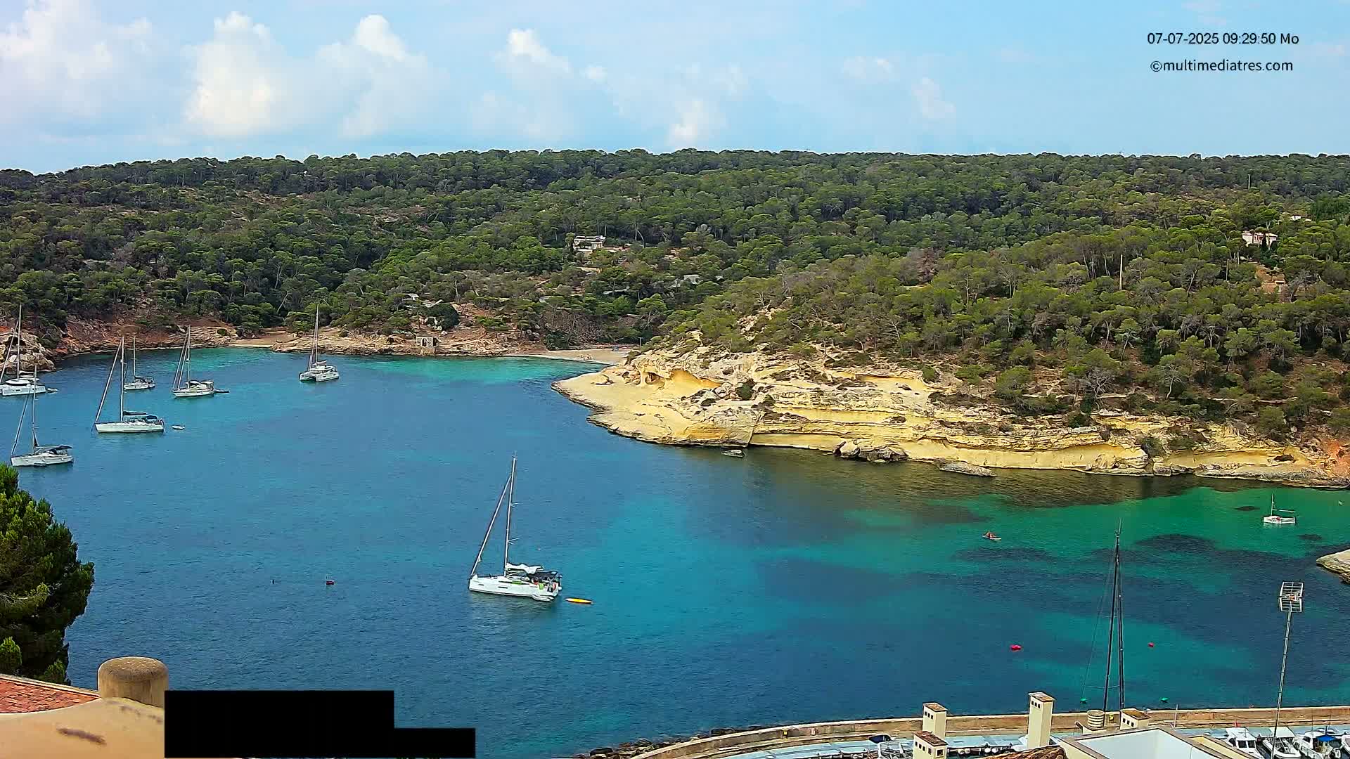 A secluded turquoise bay is surrounded by a lush green, hilly landscape under a partly cloudy sky, with several sailboats anchored in the calm water.