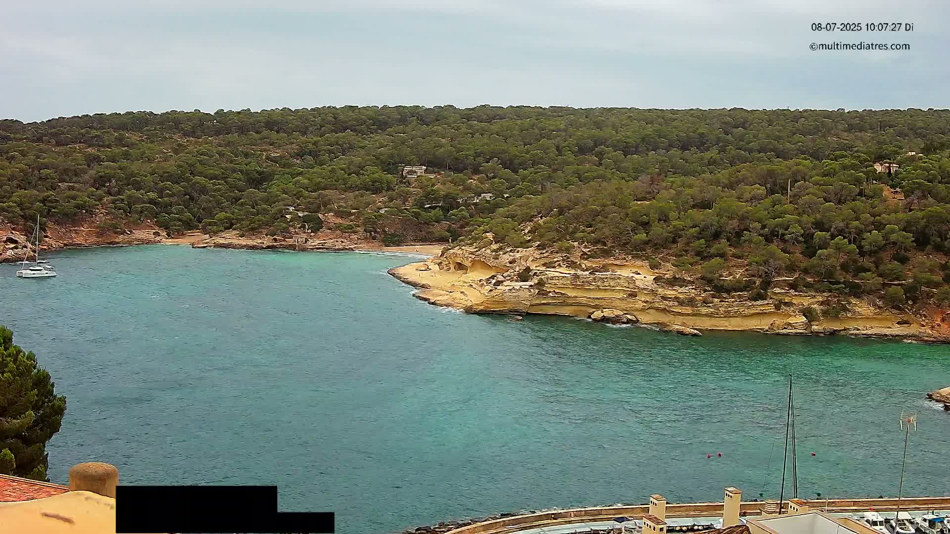 A secluded cove with turquoise water is surrounded by a lush, green hillside under a cloudy sky.